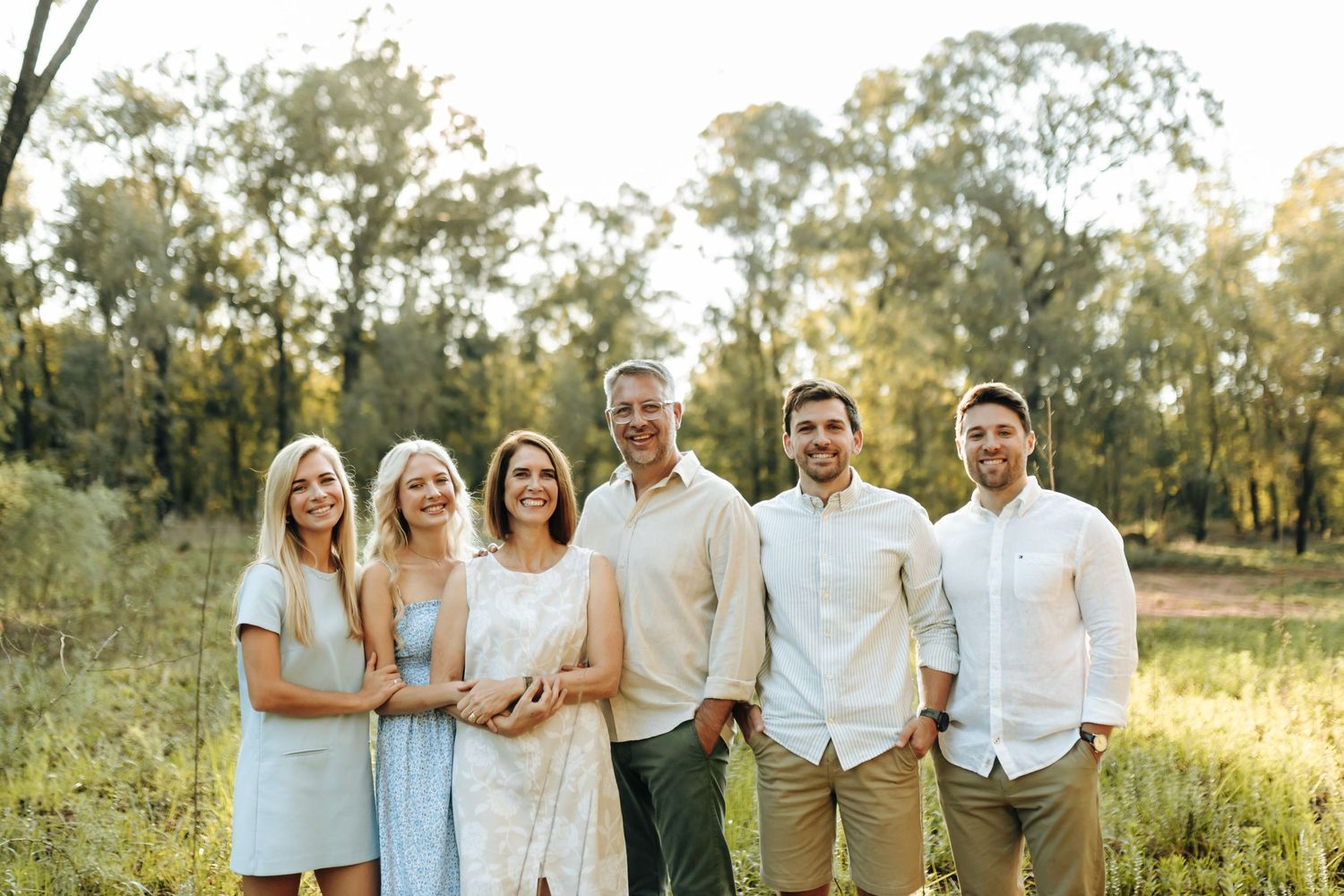 A group of six people in light colored clothing standing together in a sunny field with trees in the background.