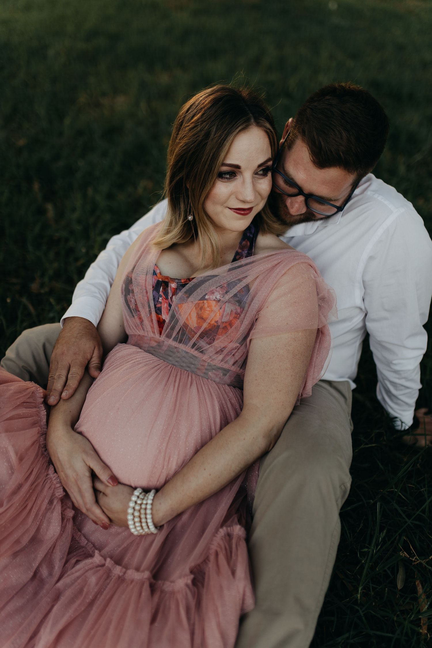 A couple sits together outdoors during a maternity photo shoot, with the woman wearing a flowing pink dress and pearl bracelet.