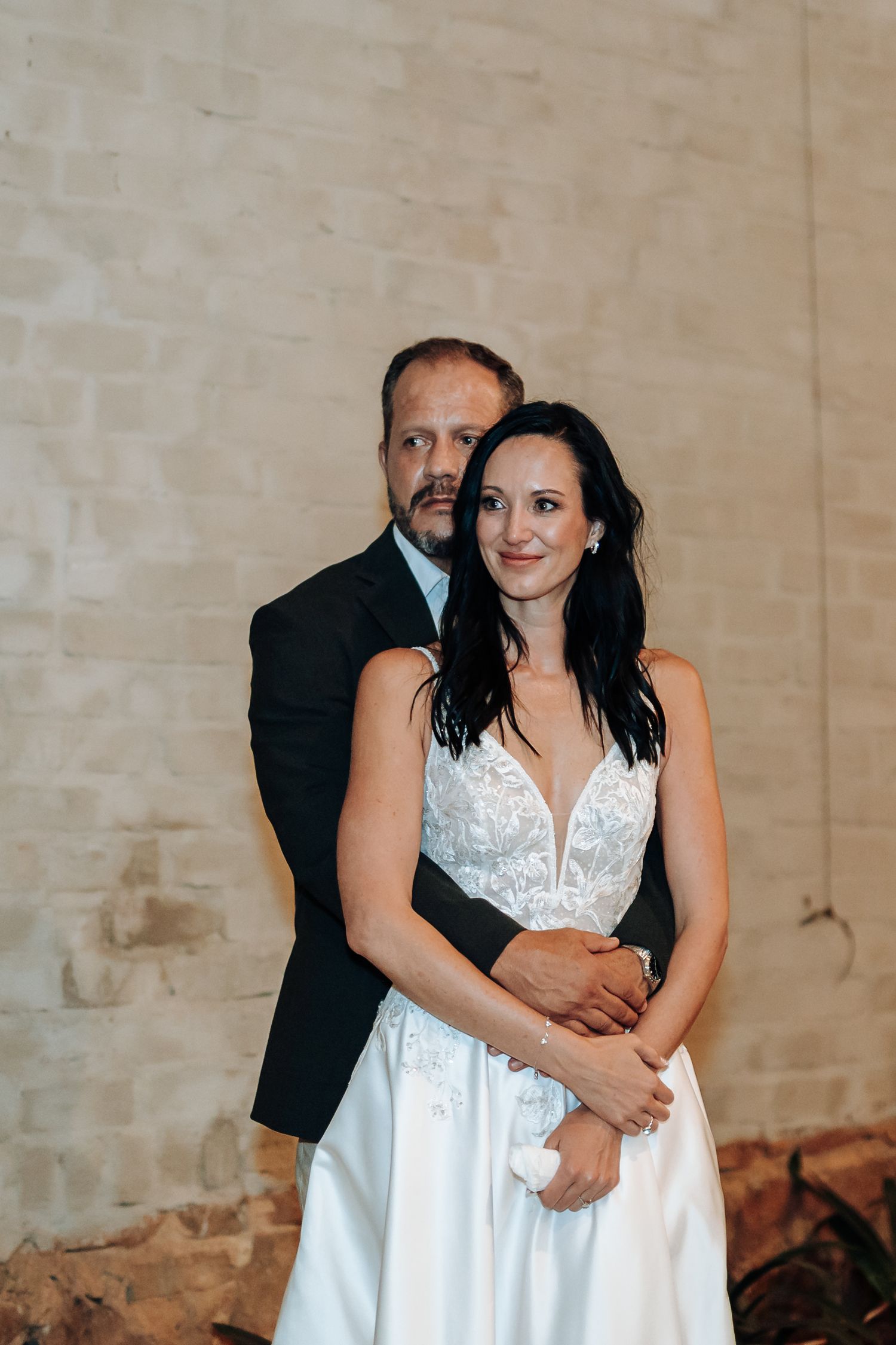 Bride in white lace gown embraced by groom in black suit, posing before rustic stone wall.