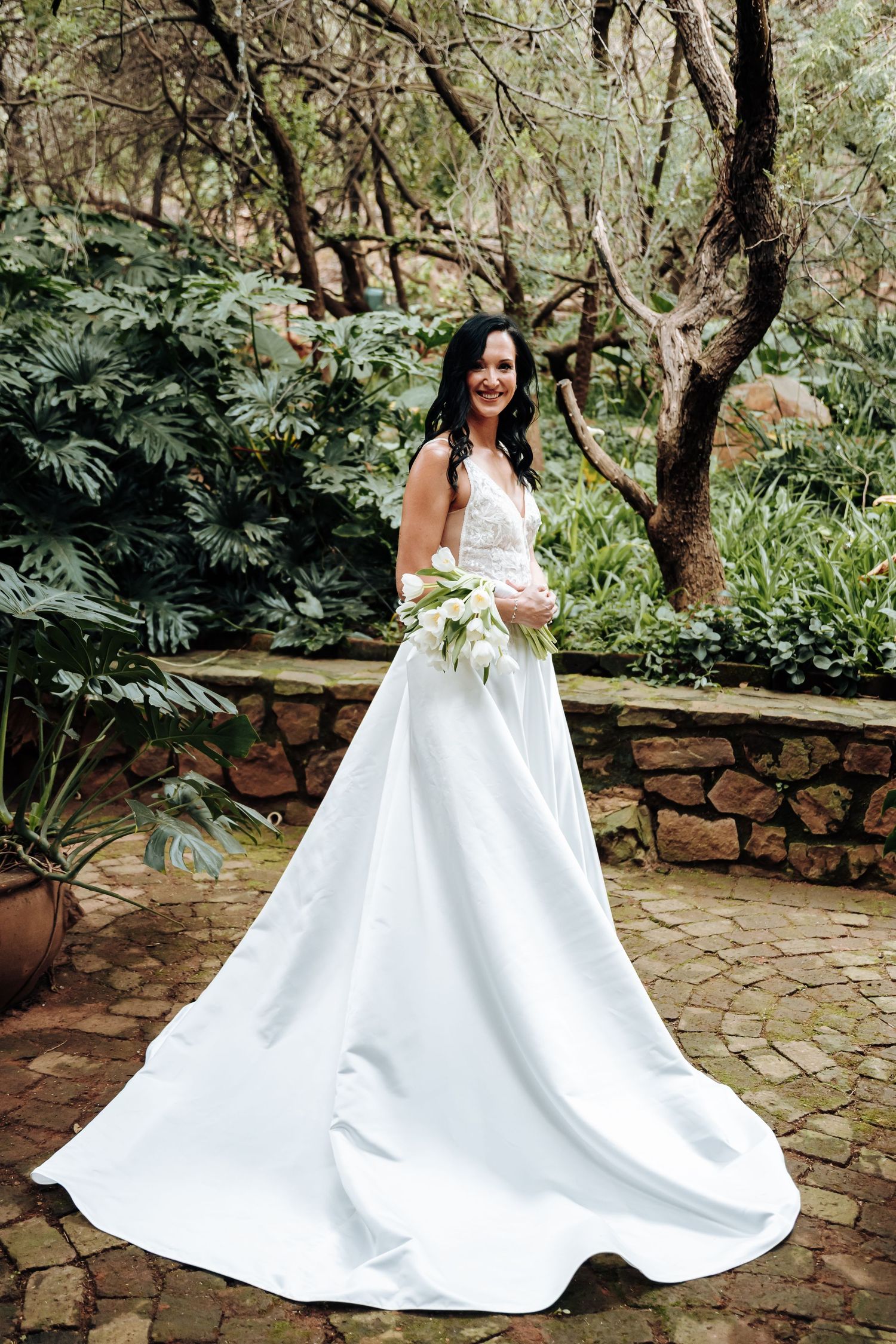 Bride in elegant white ball gown with train holds bouquet in lush garden setting.