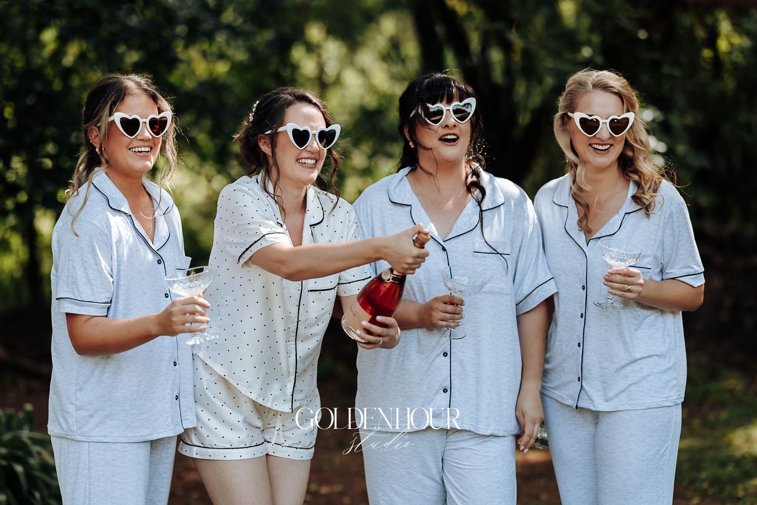Four bridesmaids in matching light blue pajamas celebrate with champagne and heart-shaped sunglasses outdoors.