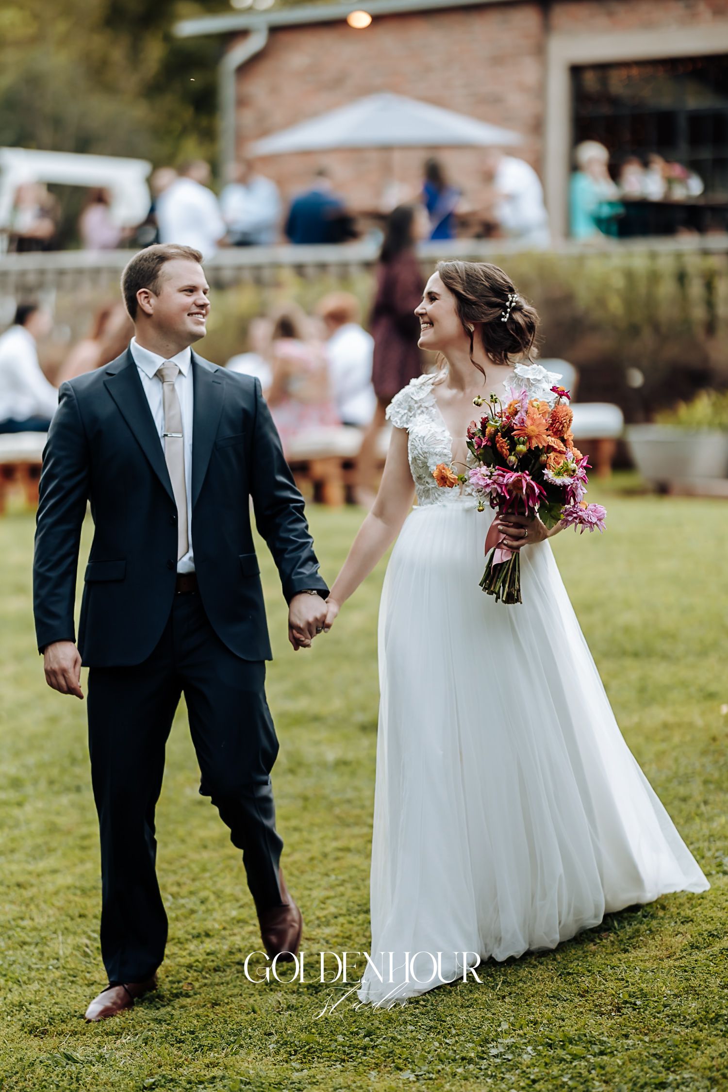 Bride and groom walk hand in hand across green lawn with colorful bridal bouquet.