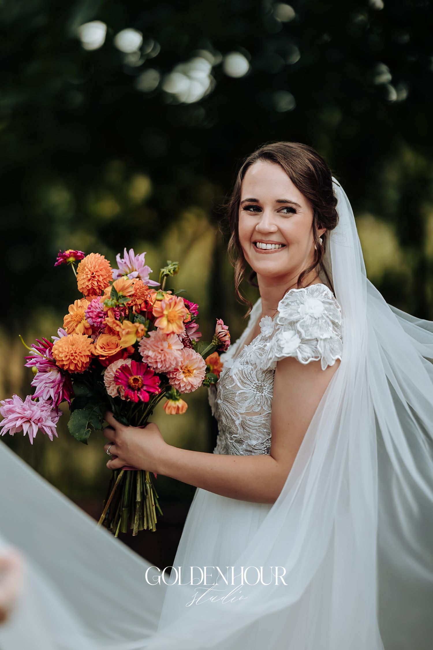 Smiling bride in lace gown holds vibrant pink and orange bouquet with flowing veil outdoors.