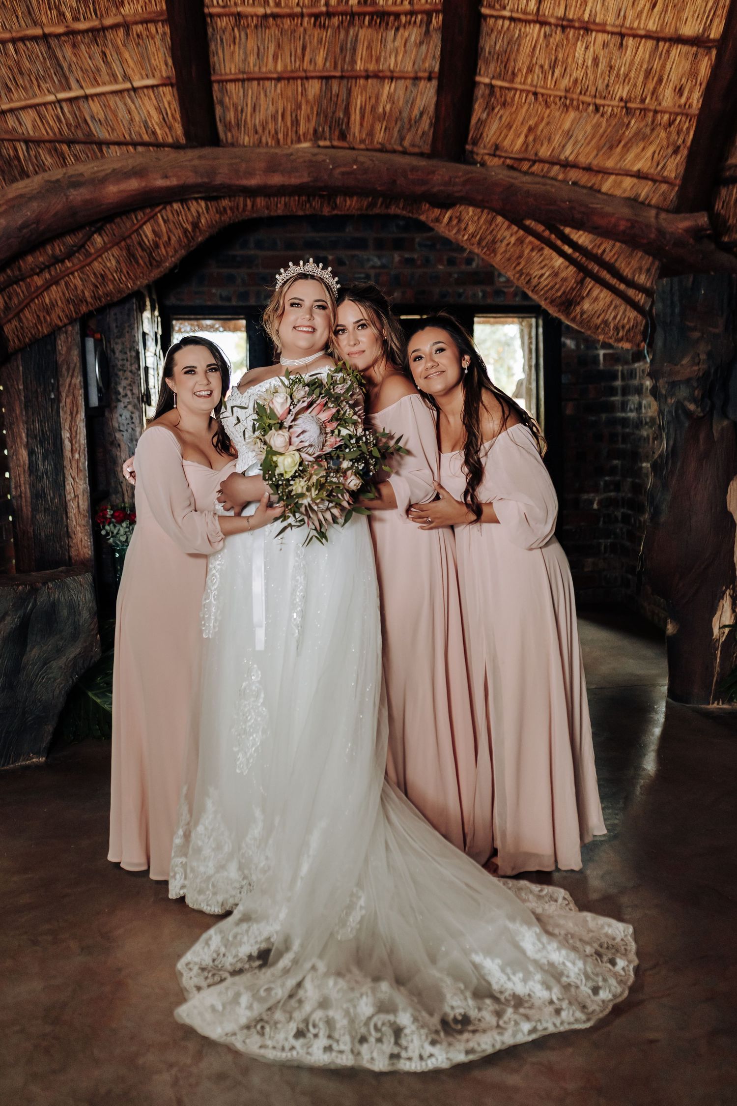 Bride in white lace gown poses with bridesmaids wearing blush pink dresses under rustic thatched roof venue.