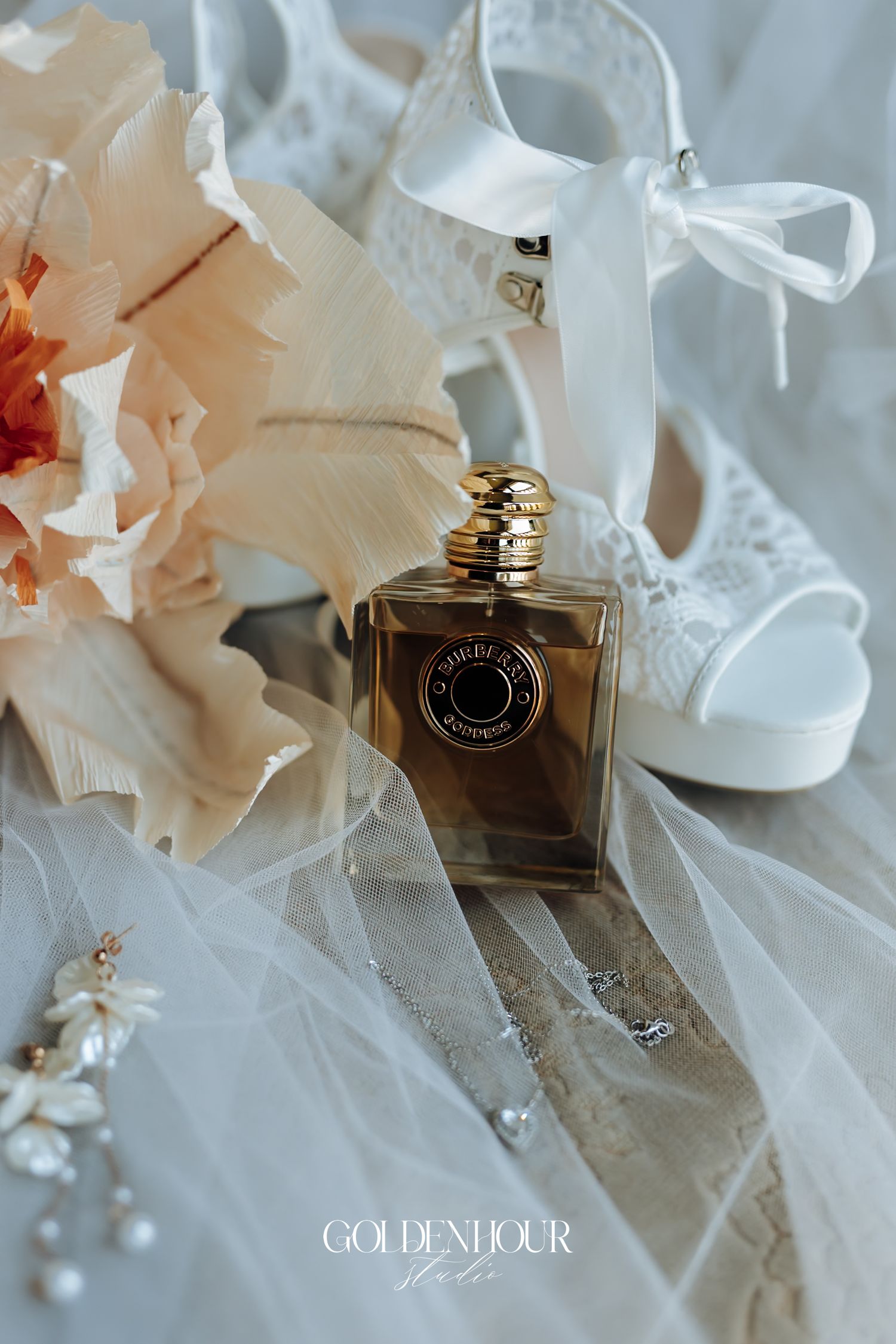 Bridal flat lay with perfume bottle, white heels, lace veil, floral bouquet, and crystal earrings on soft fabric.