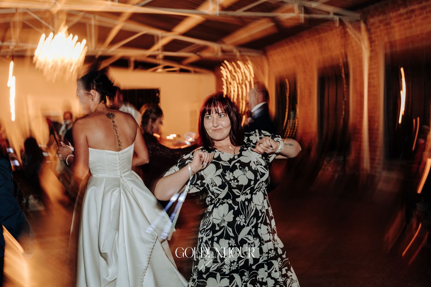 Bride in white satin gown dances joyfully with a guest in a floral dress at a lively wedding reception.