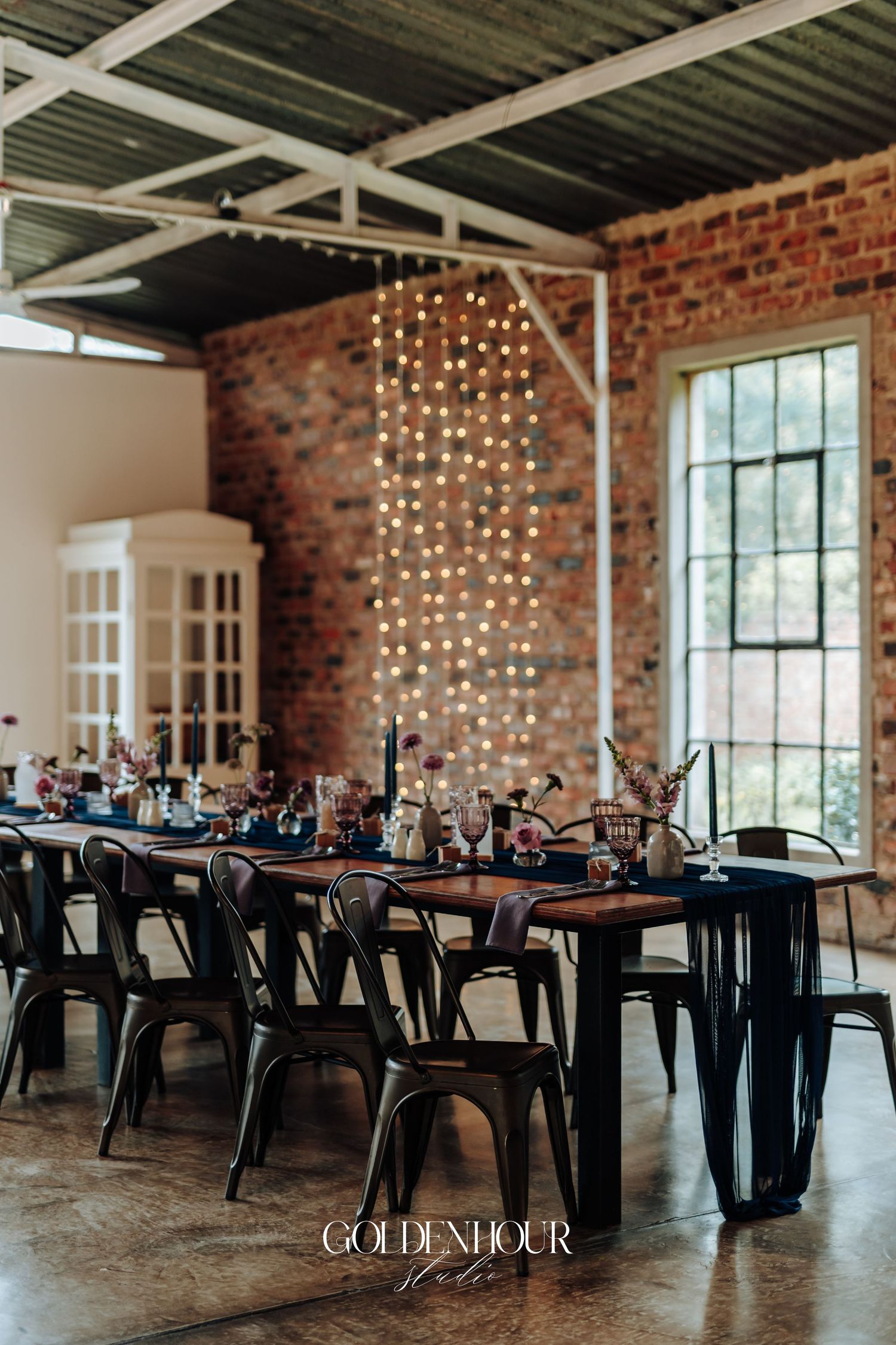 Industrial wedding reception with string lights, brick walls, dark wood tables, navy linens, and metal chairs in an elegant venue.