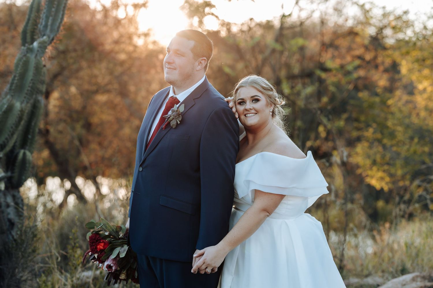 A bride and groom hold hands outdoors at golden hour, with cacti and autumn foliage in a desert wedding setting.