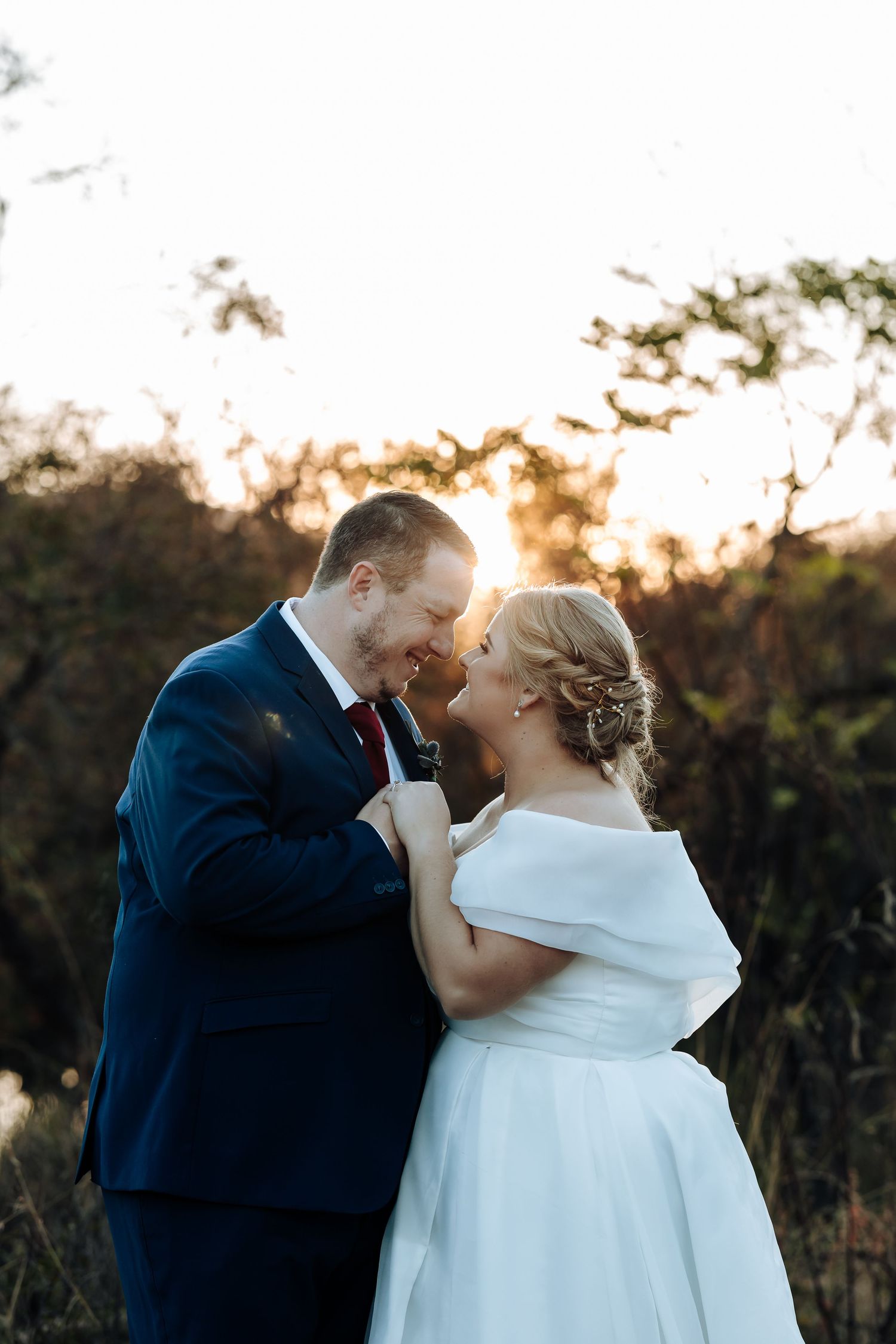 Bride and groom share a romantic moment at sunset, with golden light glowing behind them in a natural outdoor setting.