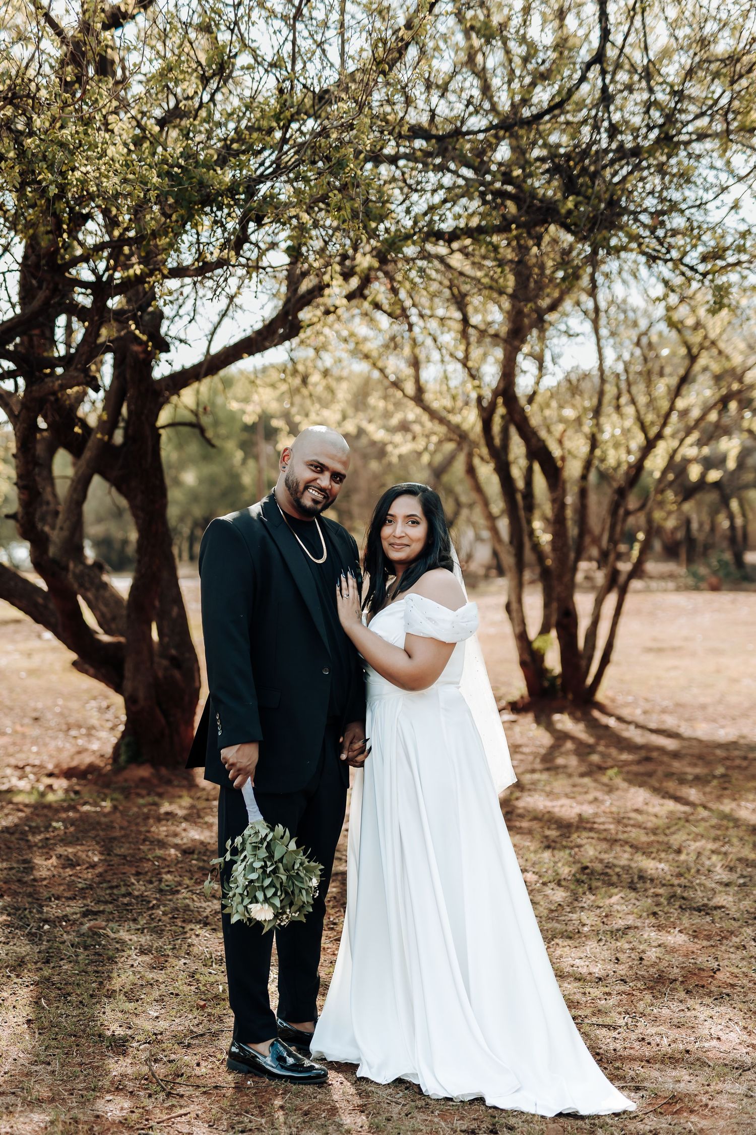 A couple in wedding attire pose together under trees in a natural outdoor setting.