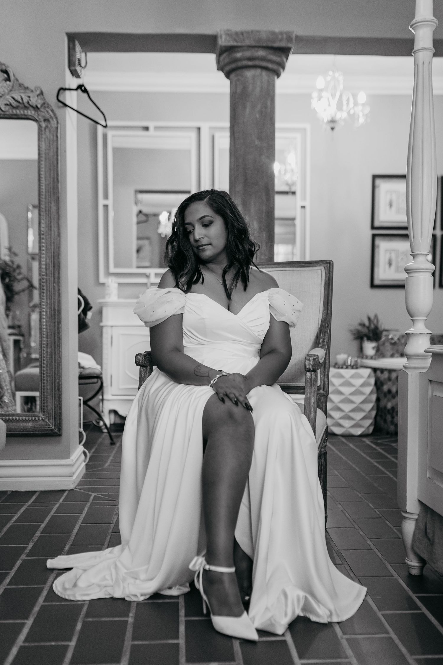 A black and white portrait of an elegant bride in a white off-shoulder gown sitting on a chair in an ornate room.