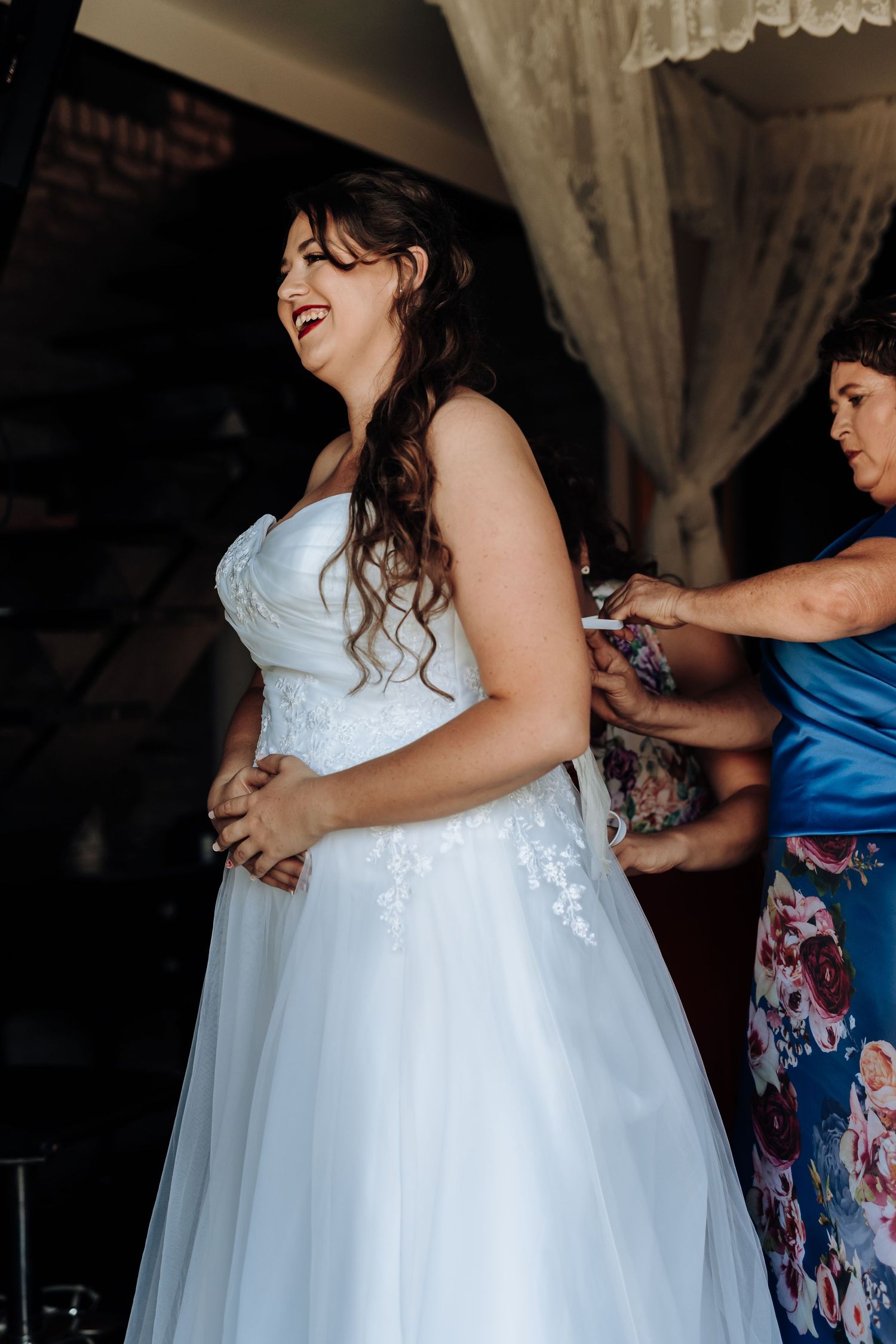 A bride in a white lace wedding dress laughs joyfully while being helped into her gown.