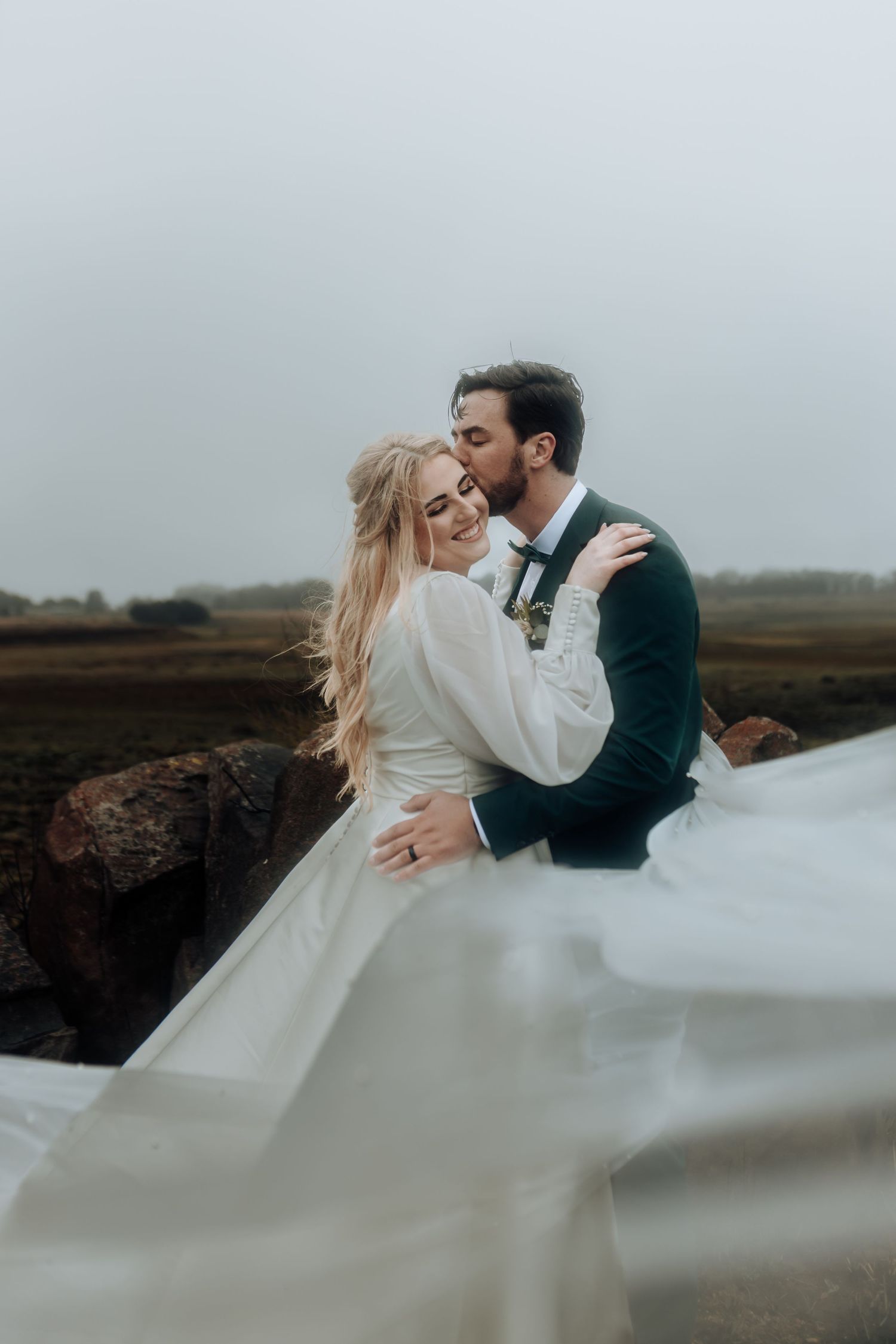 Newlyweds share an intimate moment against misty backdrop as bride's dress catches the wind.