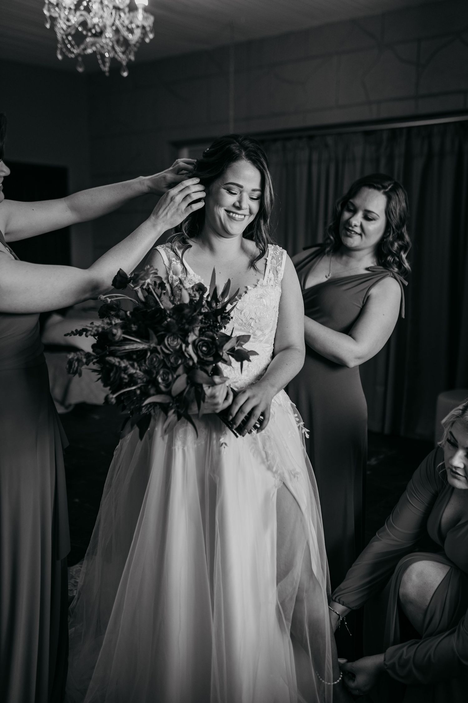 Black and white photo of bride holding bouquet while getting final touches from wedding party.
