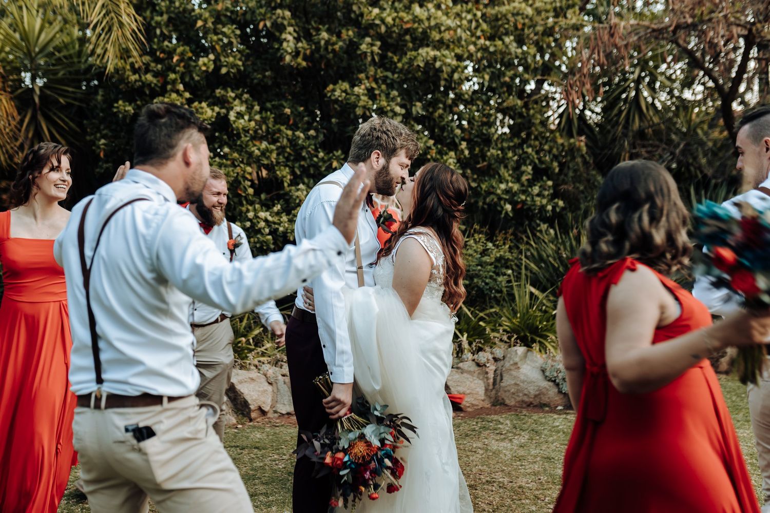 Wedding celebration in garden setting with guests in red dresses and groomsmen in suspenders.