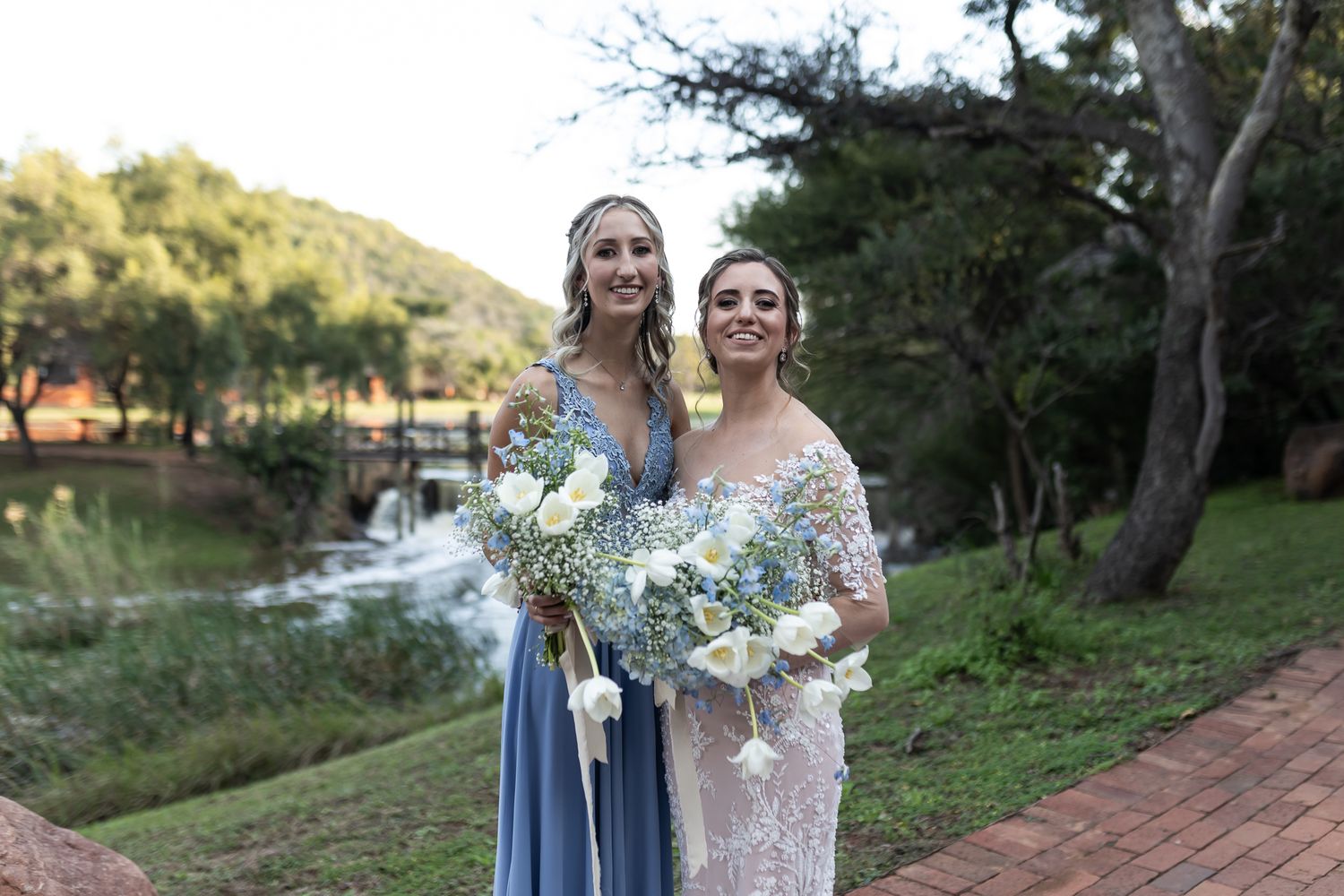 Two women pose with bouquets against natural backdrop during outdoor wedding celebration.