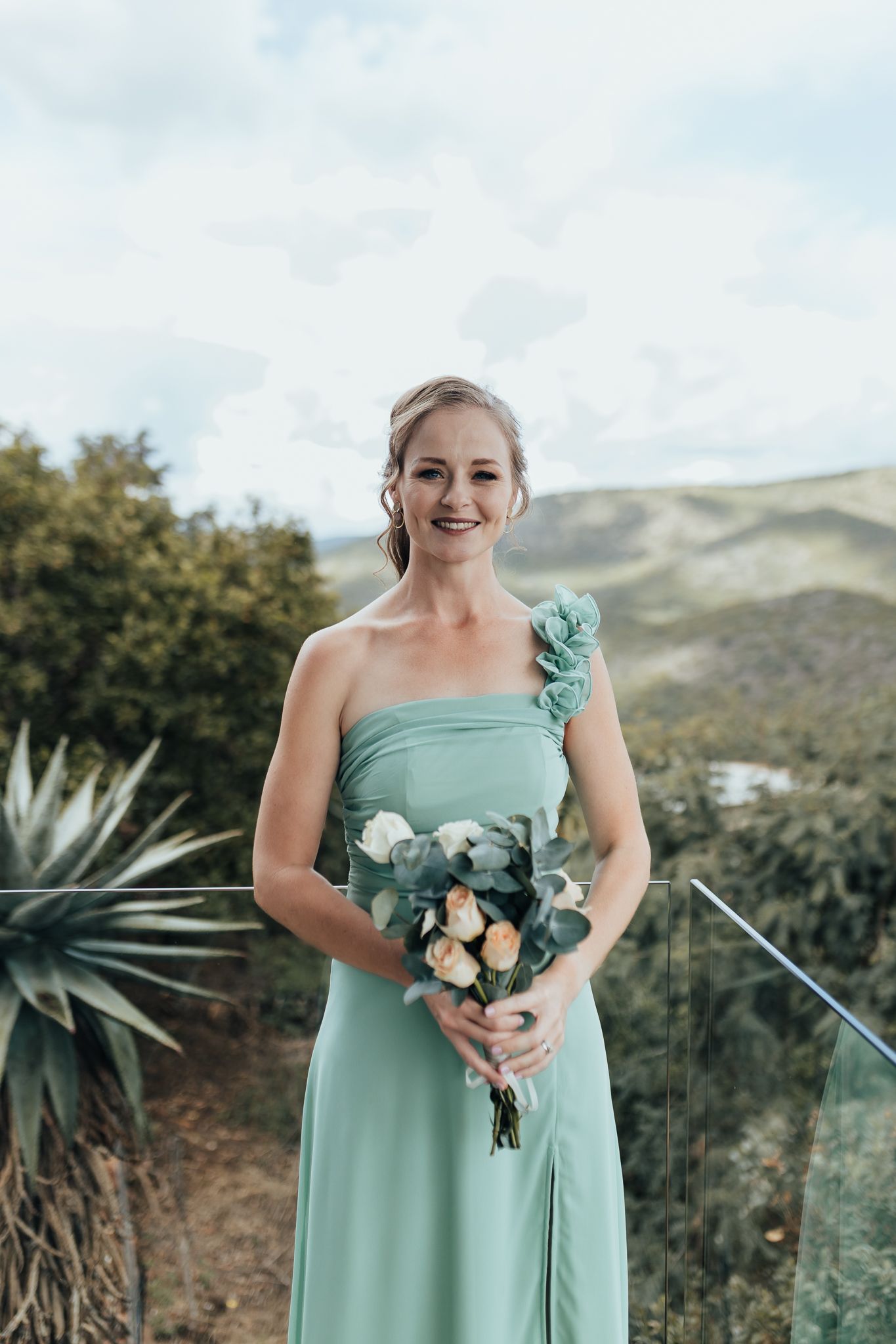Bridesmaid in mint green dress holds a matching floral bouquet against a scenic mountain landscape.