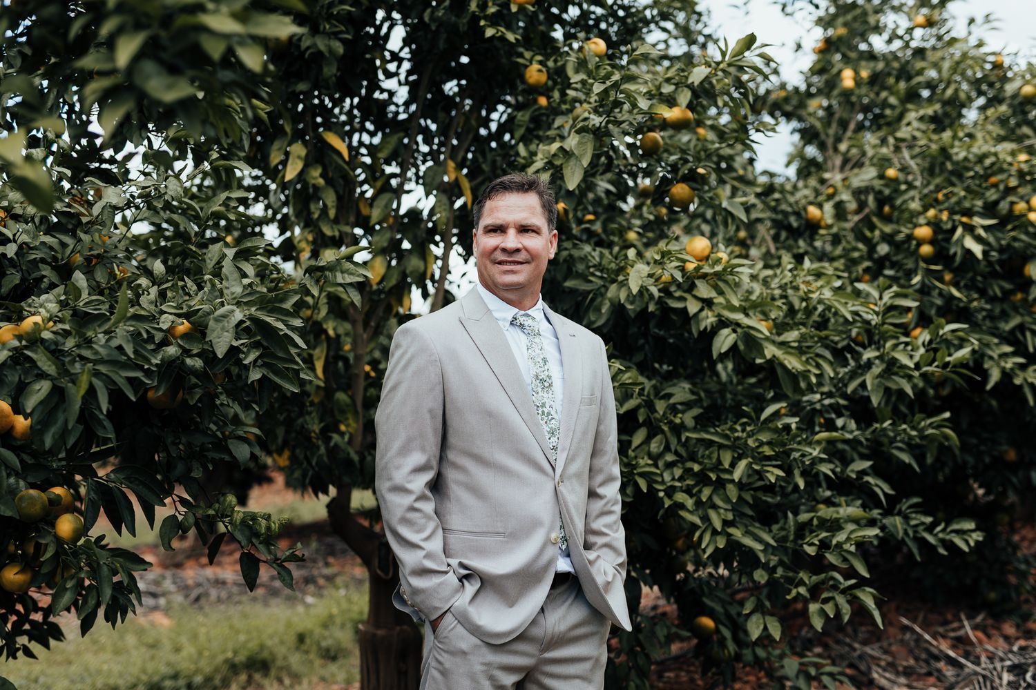 Groom in light gray suit poses in front of orange trees in an orchard.