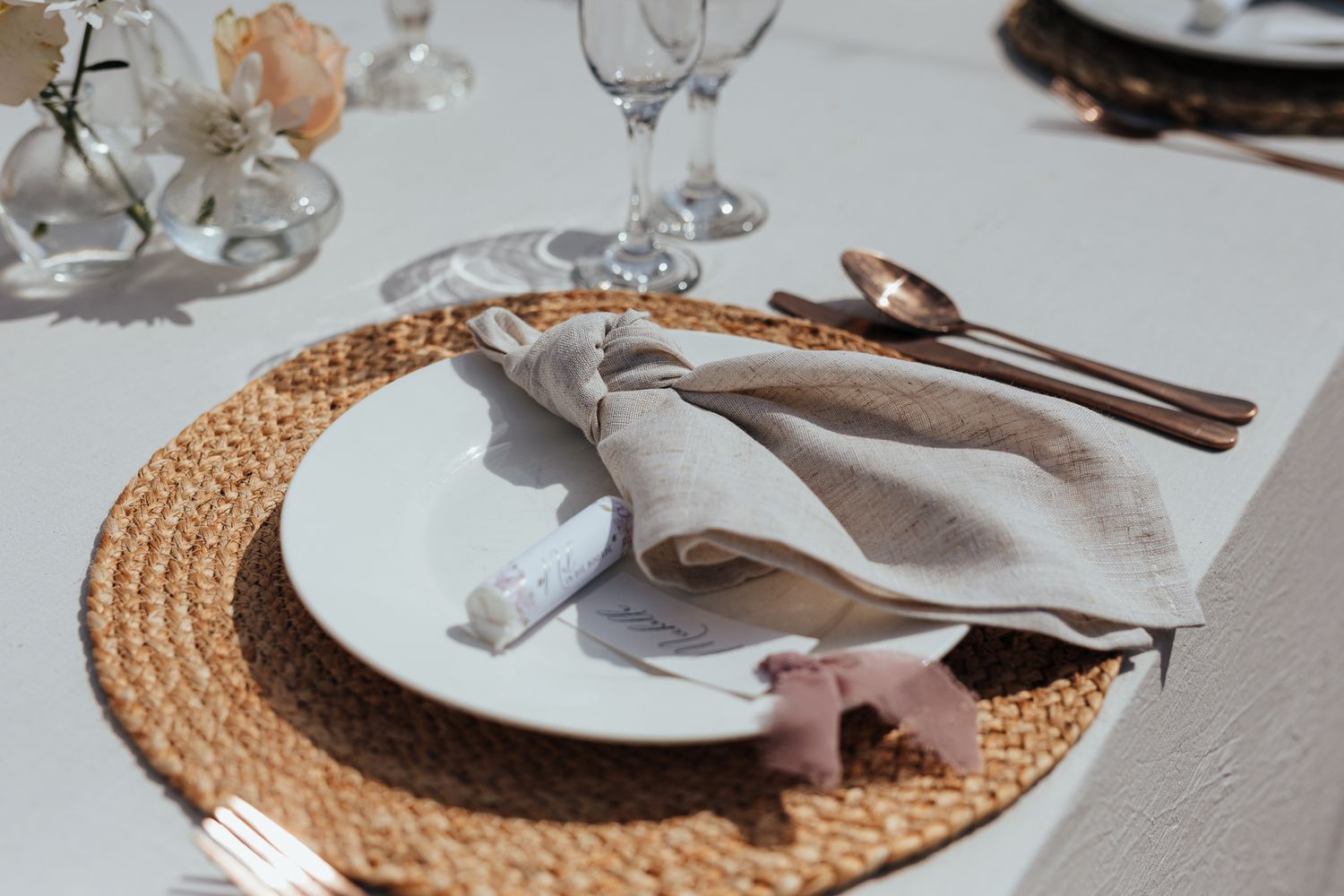 Elegant place setting with white plate on woven charger, silver cutlery, and gray linen napkin.