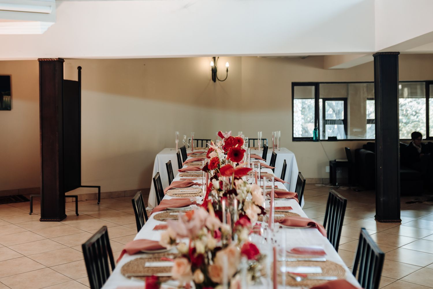 Long dining table set for wedding reception with red and pink floral arrangements and black chairs.