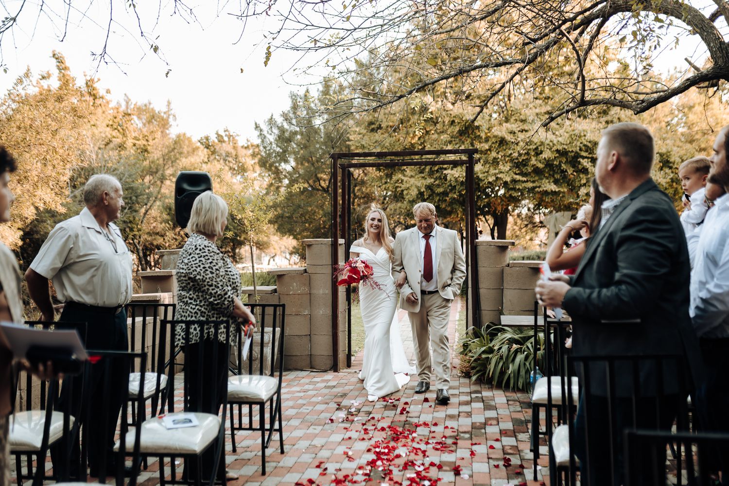 Wedding guests seated on black chairs with red rose petals lining the outdoor ceremony aisle.