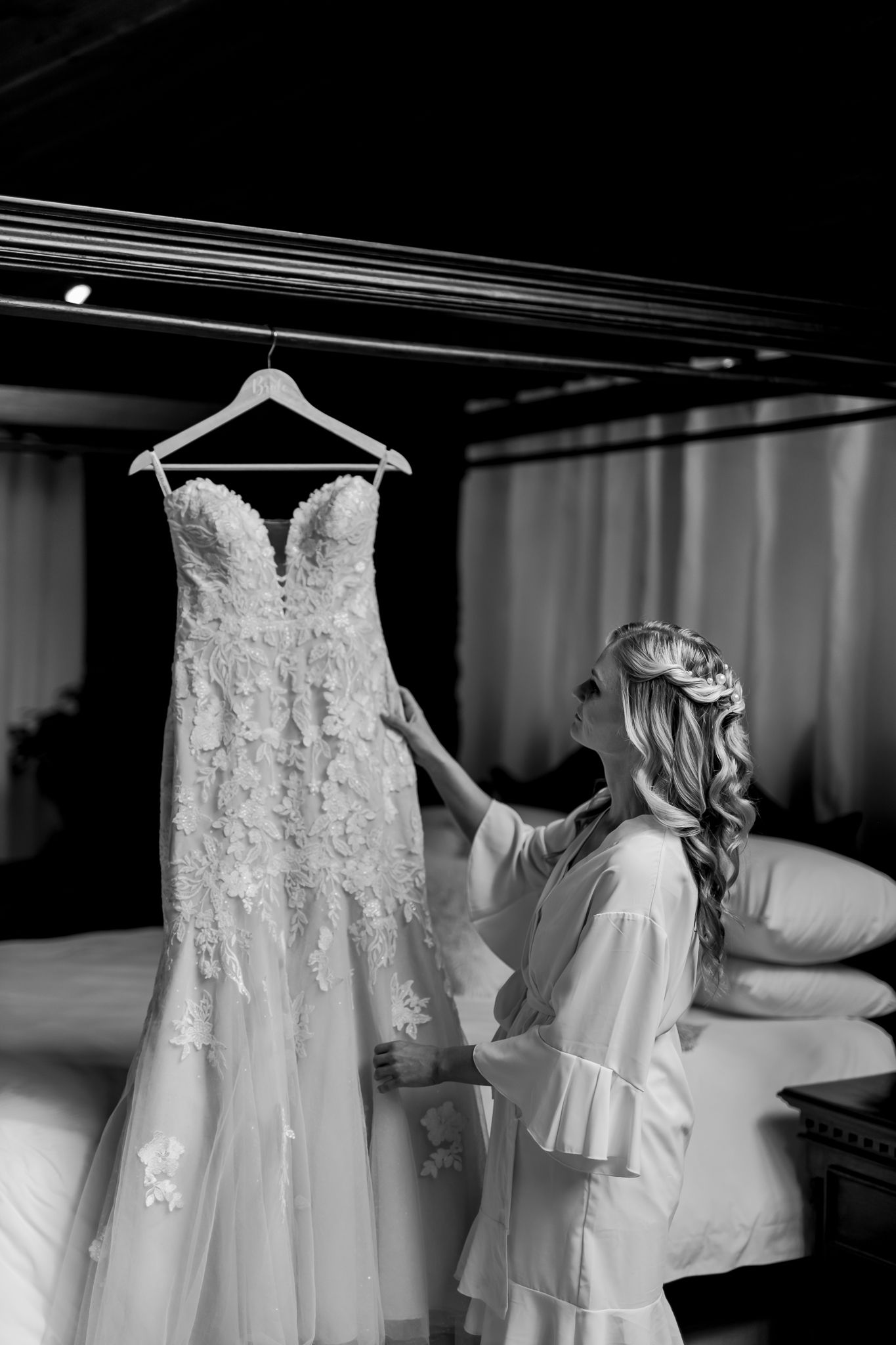 Wedding dress hangs elegantly as preparations take place in dim room setting.
