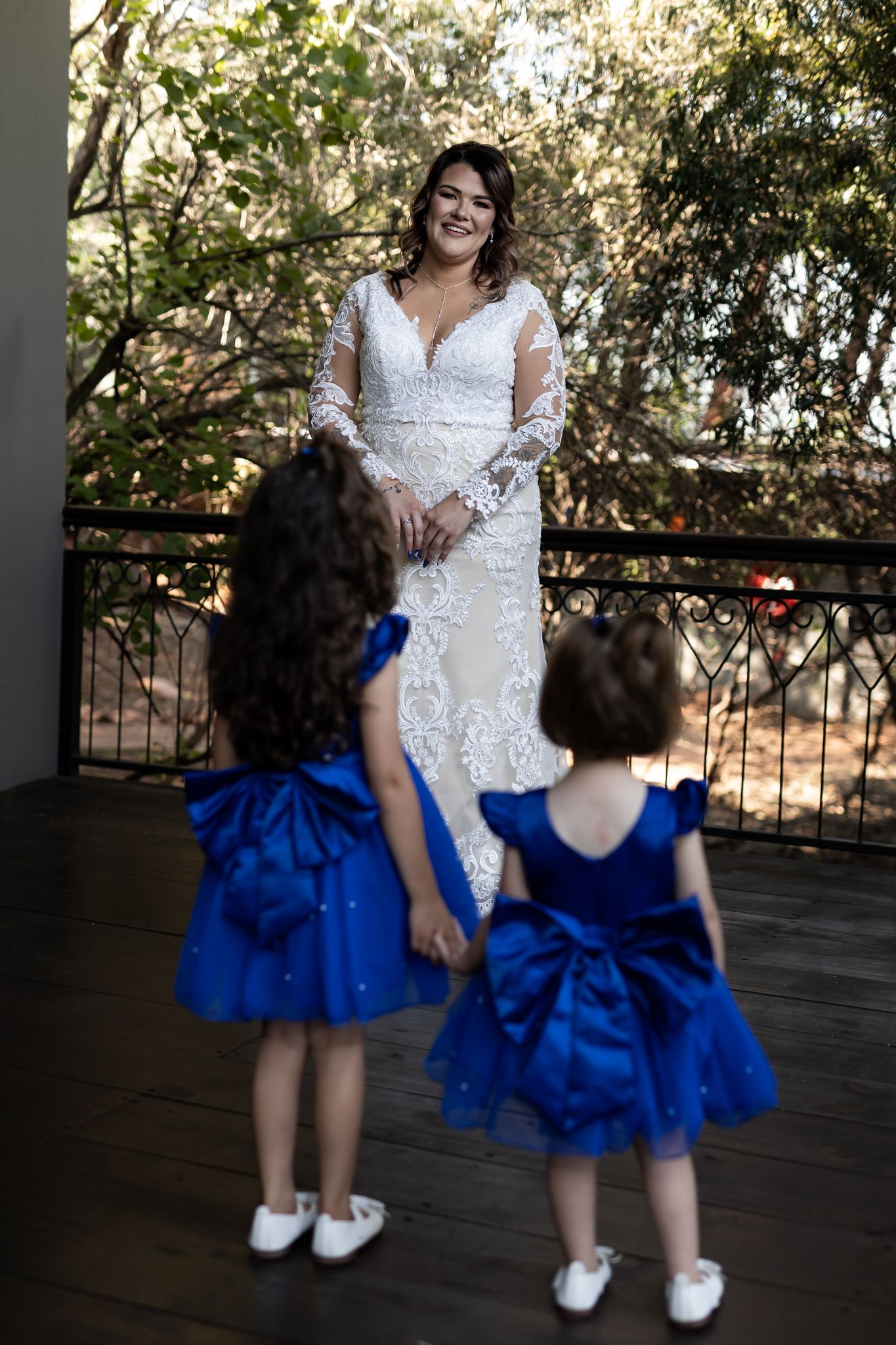 Children in bright blue dresses look up at bride in elegant lace gown on wooden deck.