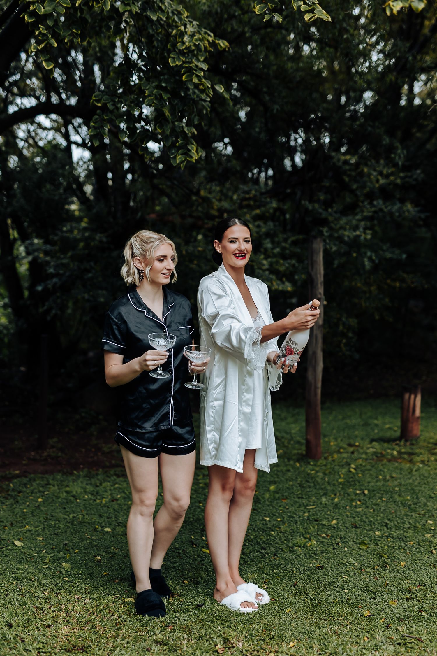 Two people in robes sharing drinks and laughing together while standing on grass near trees in an outdoor setting.