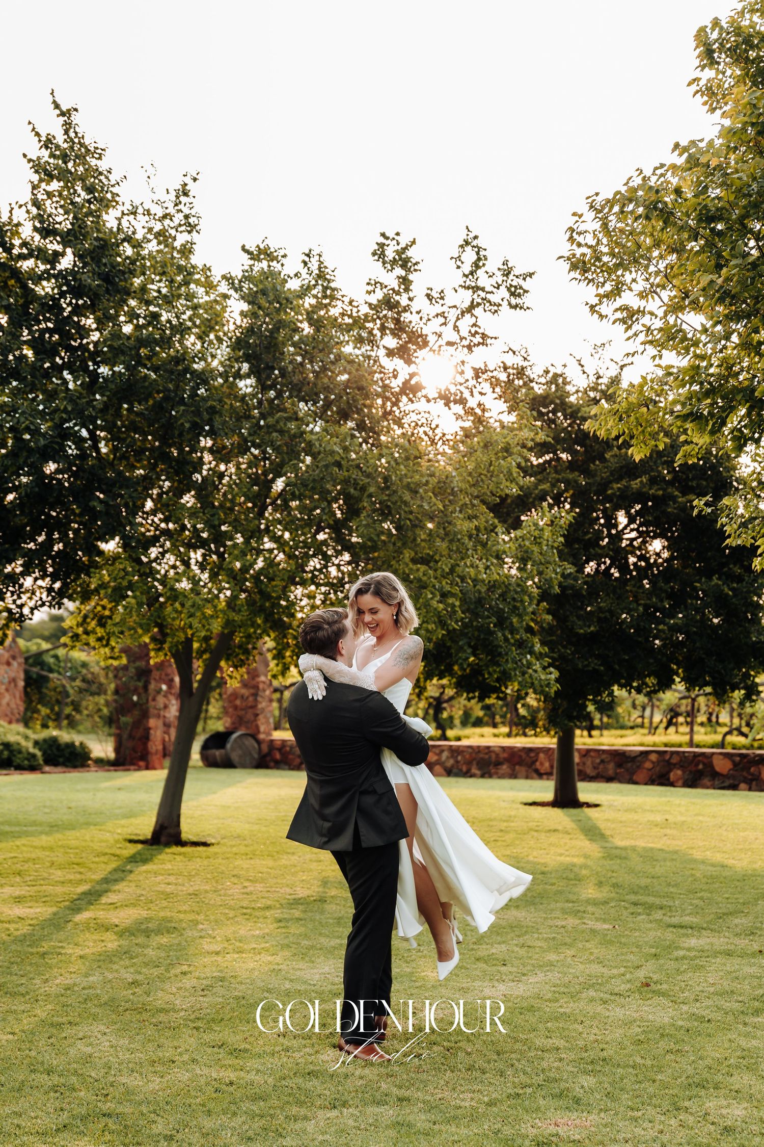 A couple dances and spins together on a grassy lawn at sunset with trees in the background.