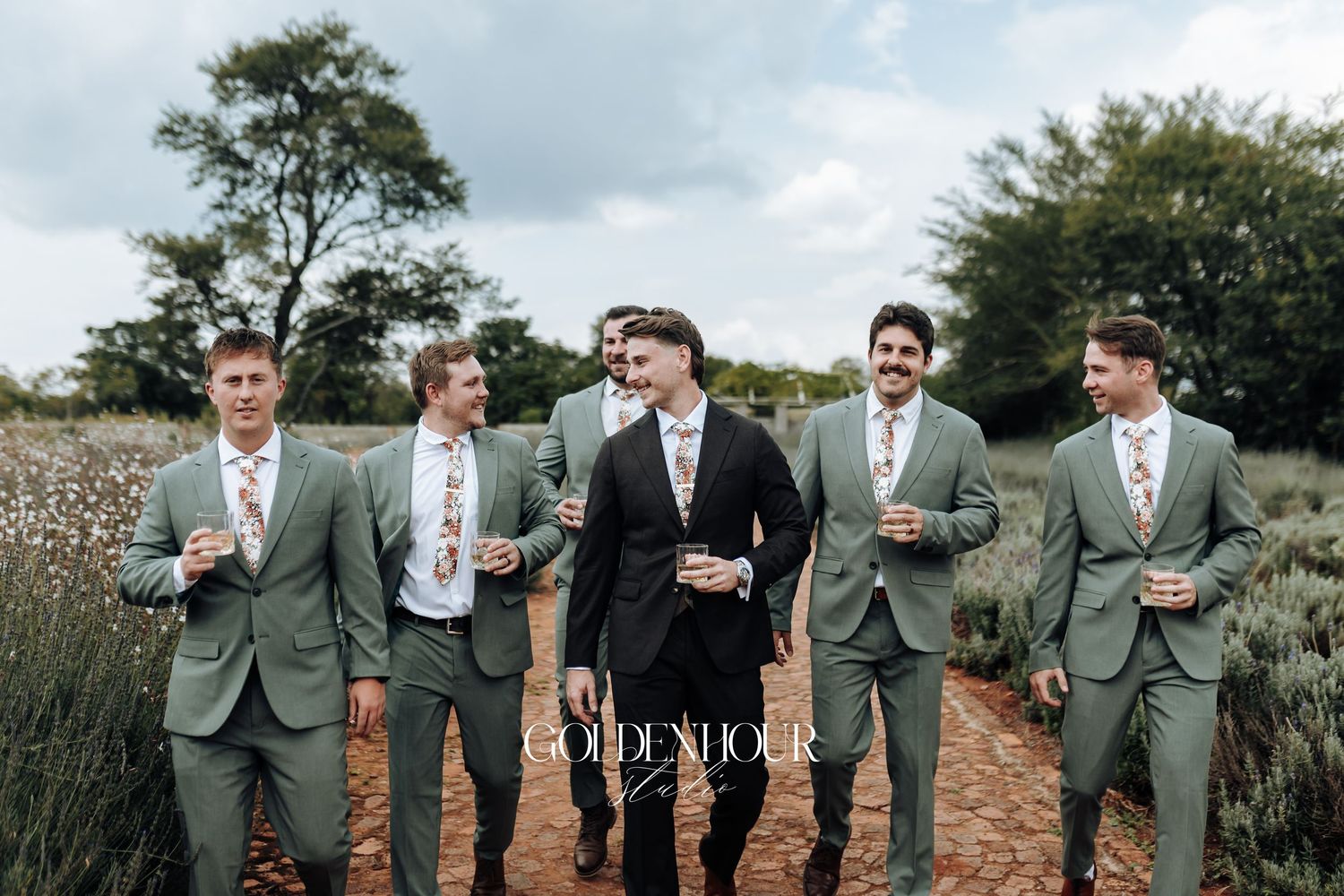 Groomsmen in gray suits share drinks and laughter during outdoor wedding celebration.