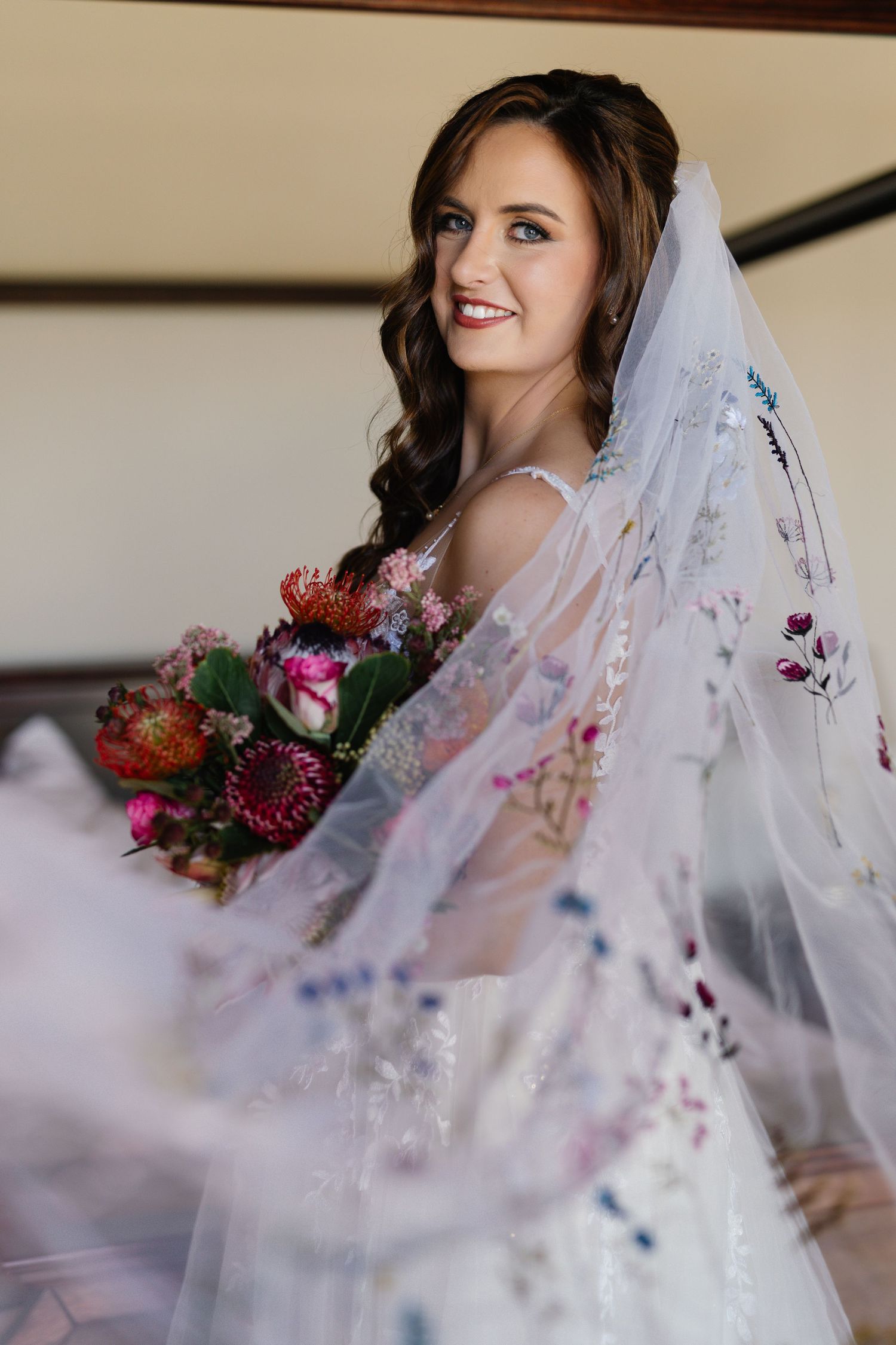 A bride in a delicate lace veil holds a vibrant burgundy and pink bouquet while getting ready for her wedding ceremony.