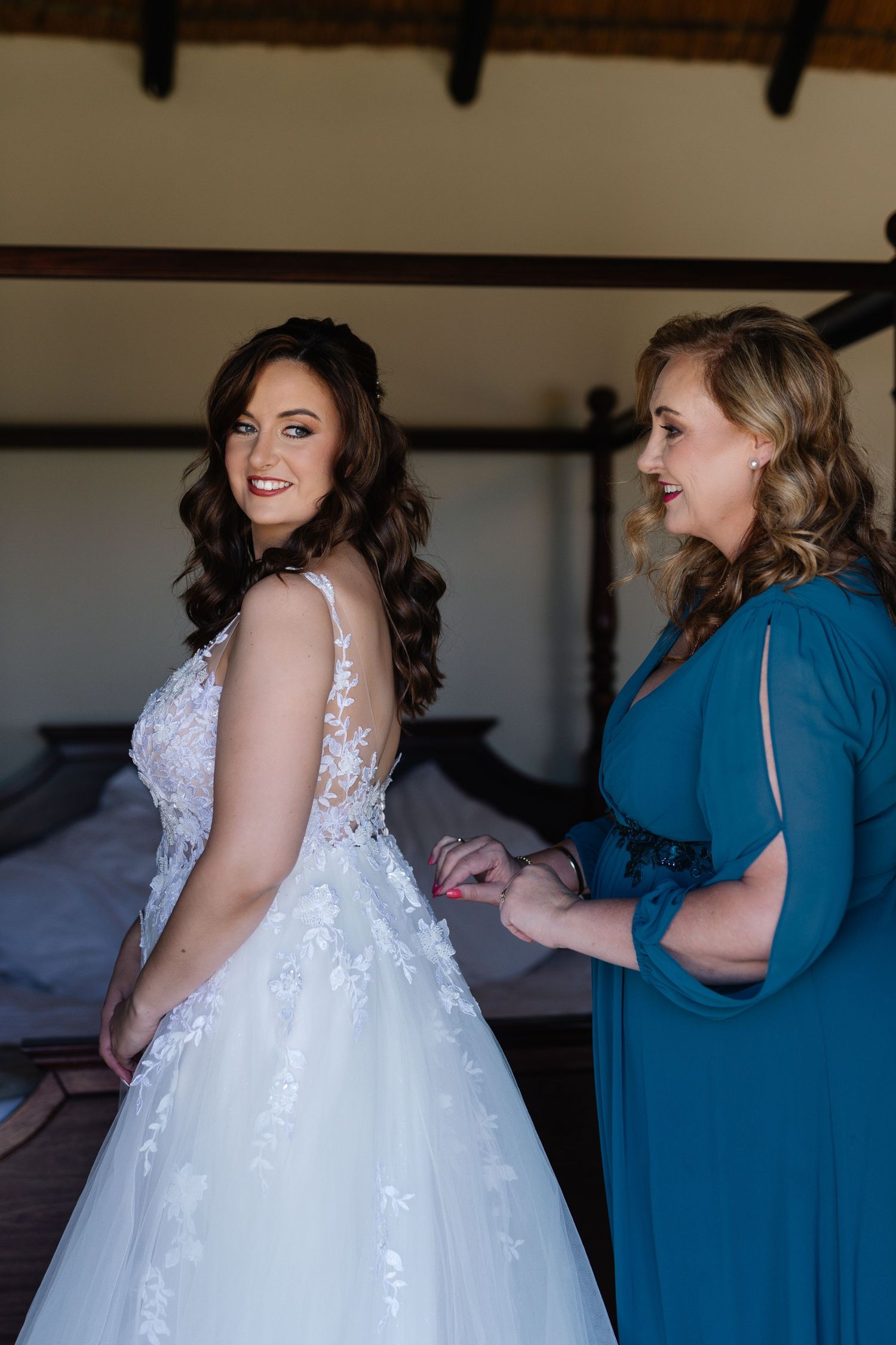 A bride in a white lace wedding gown is helped by attendants wearing teal blue dresses during wedding preparations.