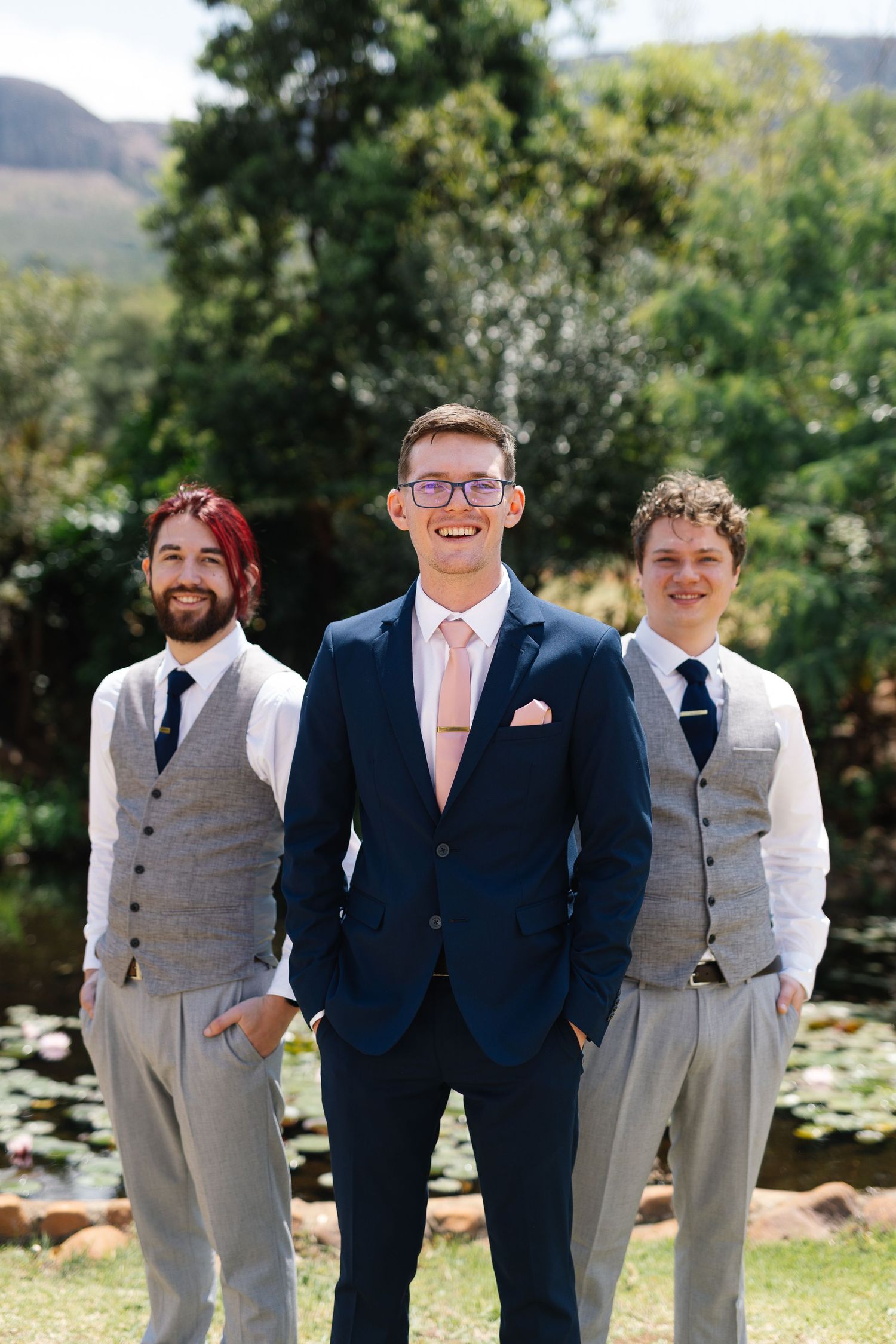 Wedding party members in navy suits and light gray vests pose together outdoors on a sunny day near mountains.