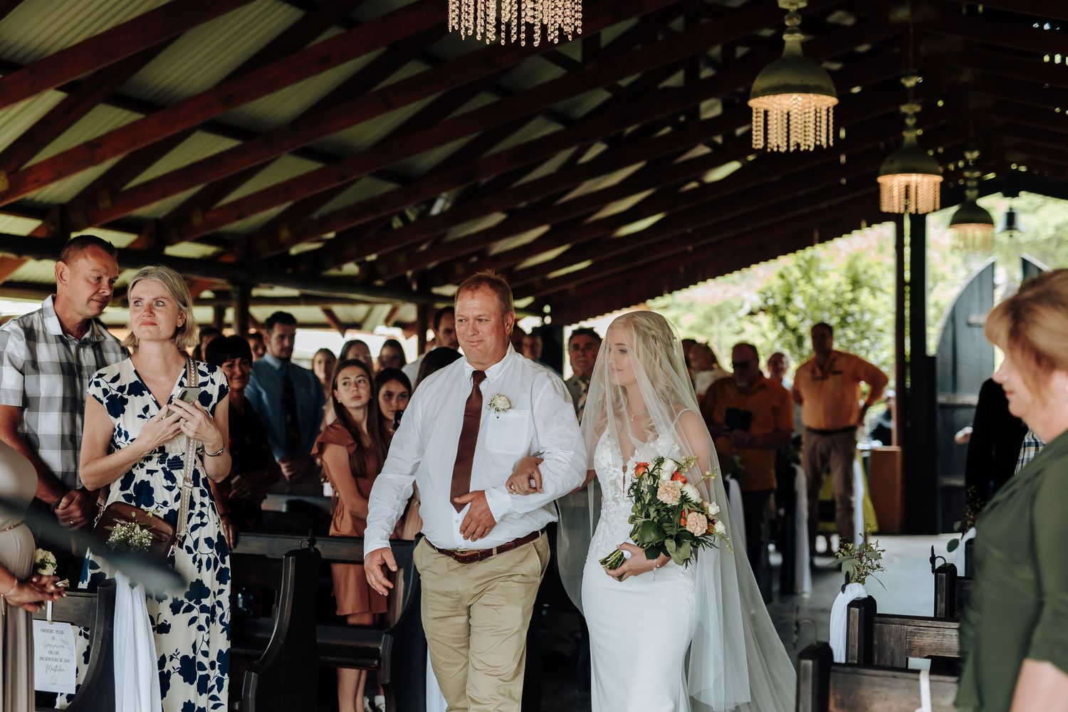 Wedding guests gather under a rustic wooden pavilion decorated with string lights and chandeliers during a celebration.