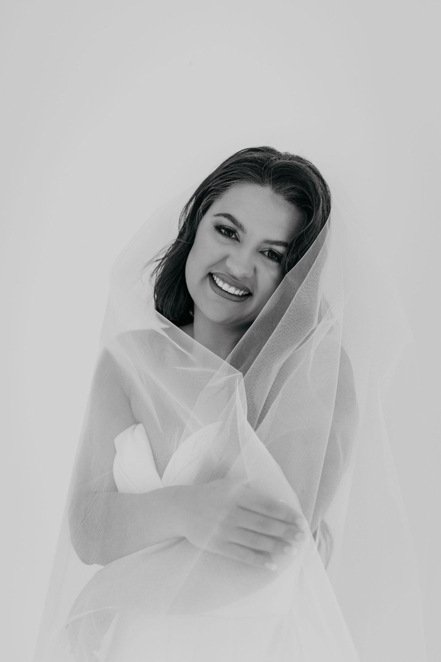 A bride wrapped in a flowing white veil smiles softly in a series of black and white portraits.