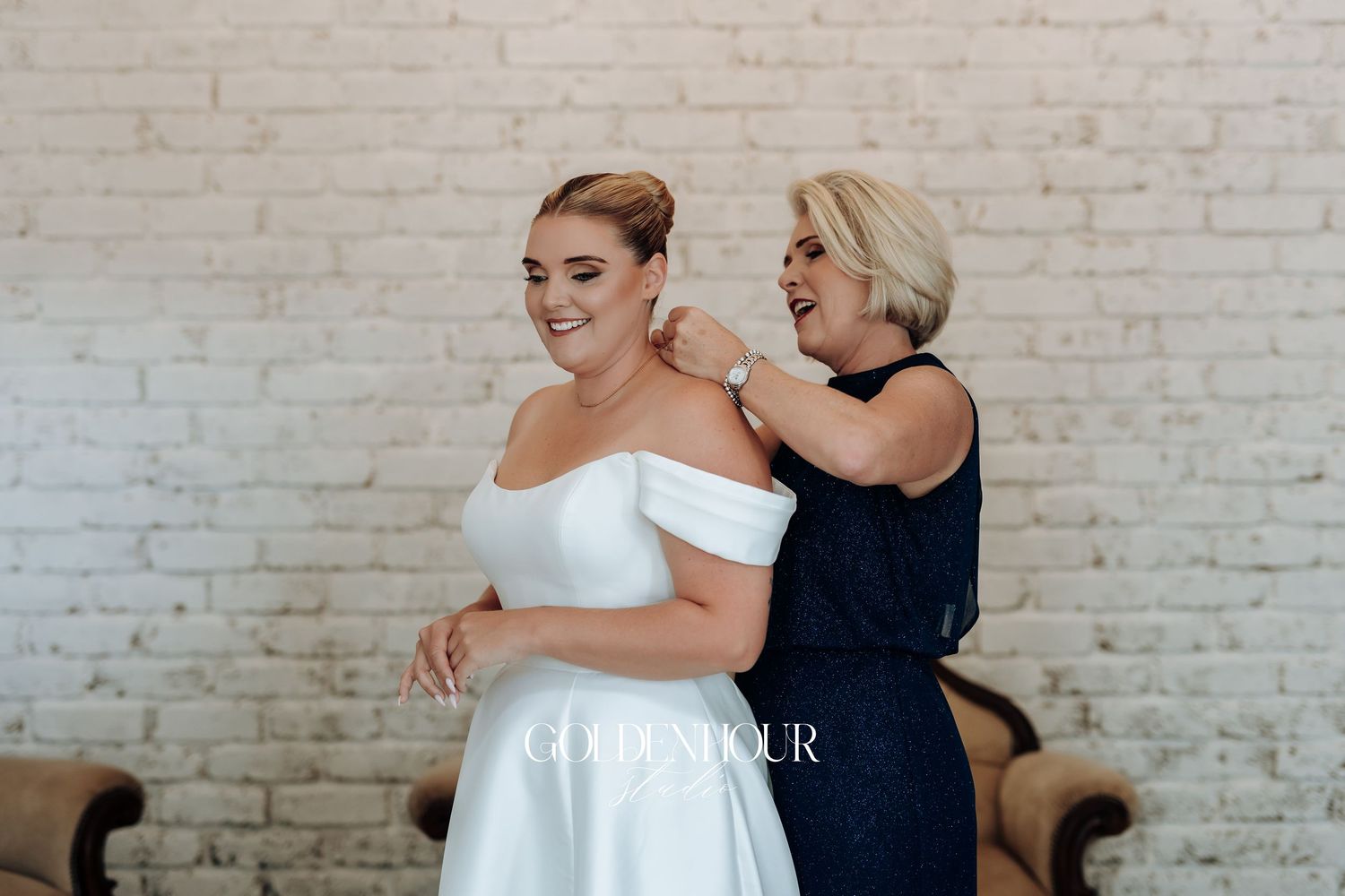 A bride in an elegant off-shoulder white gown gets help with final adjustments against a white brick wall.