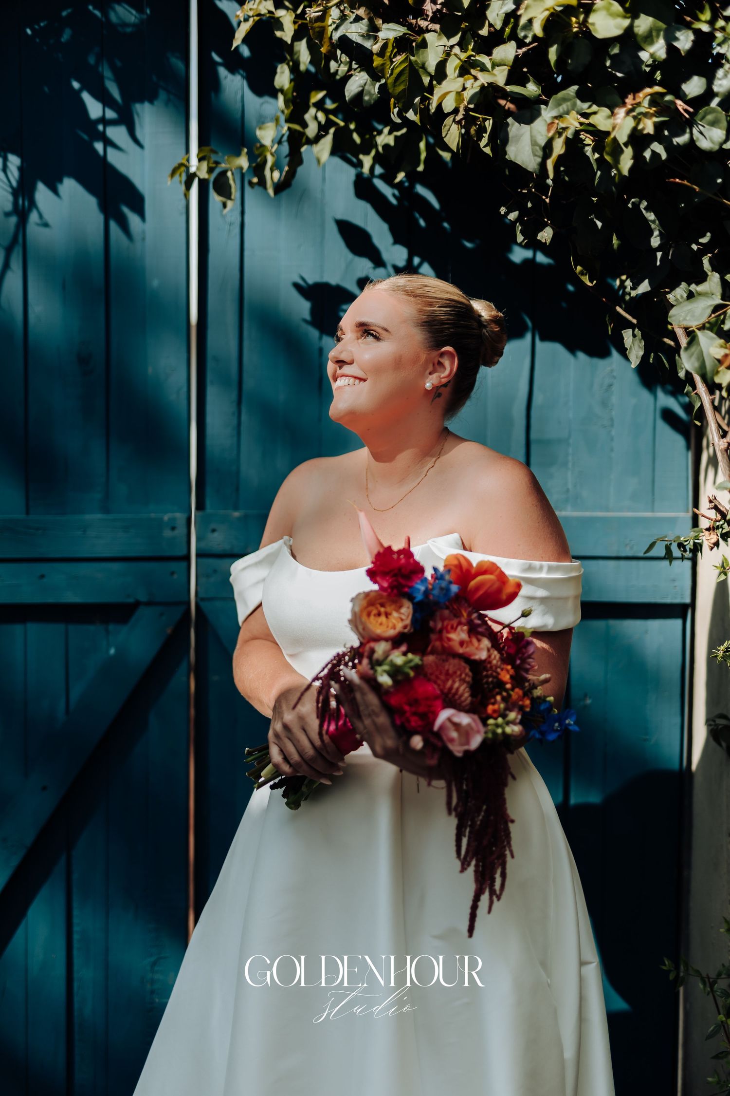 A bride in a modern white wedding dress holds a vibrant burgundy and red bouquet against a dark teal wall.