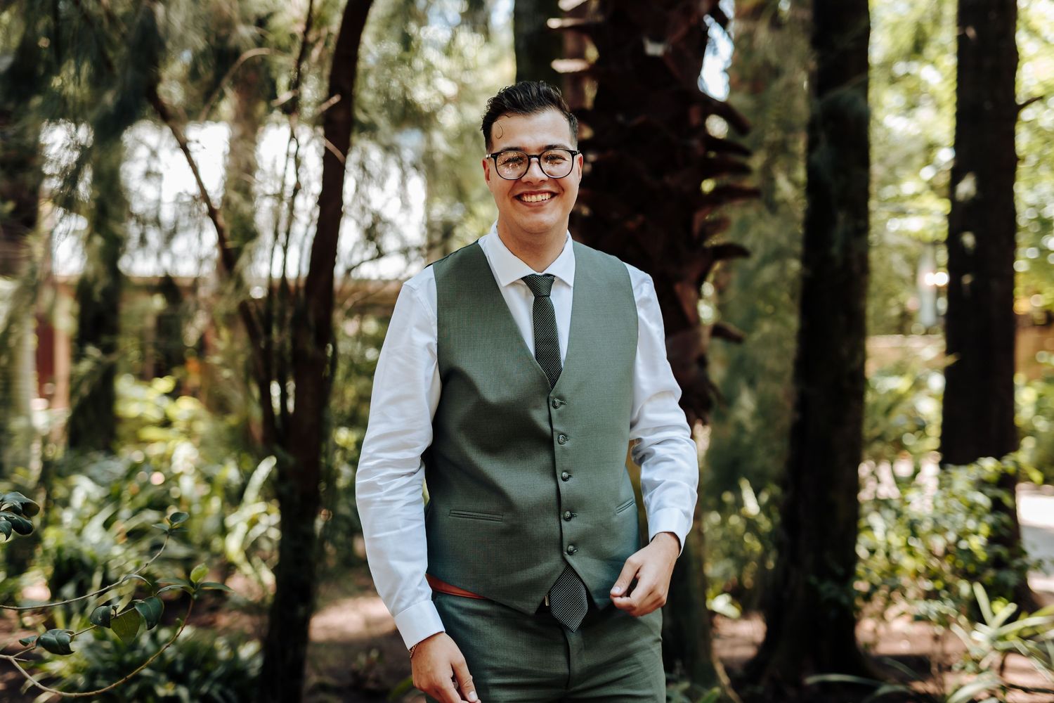 A person in a green vest and glasses smiles while standing in a sunlit forest setting.