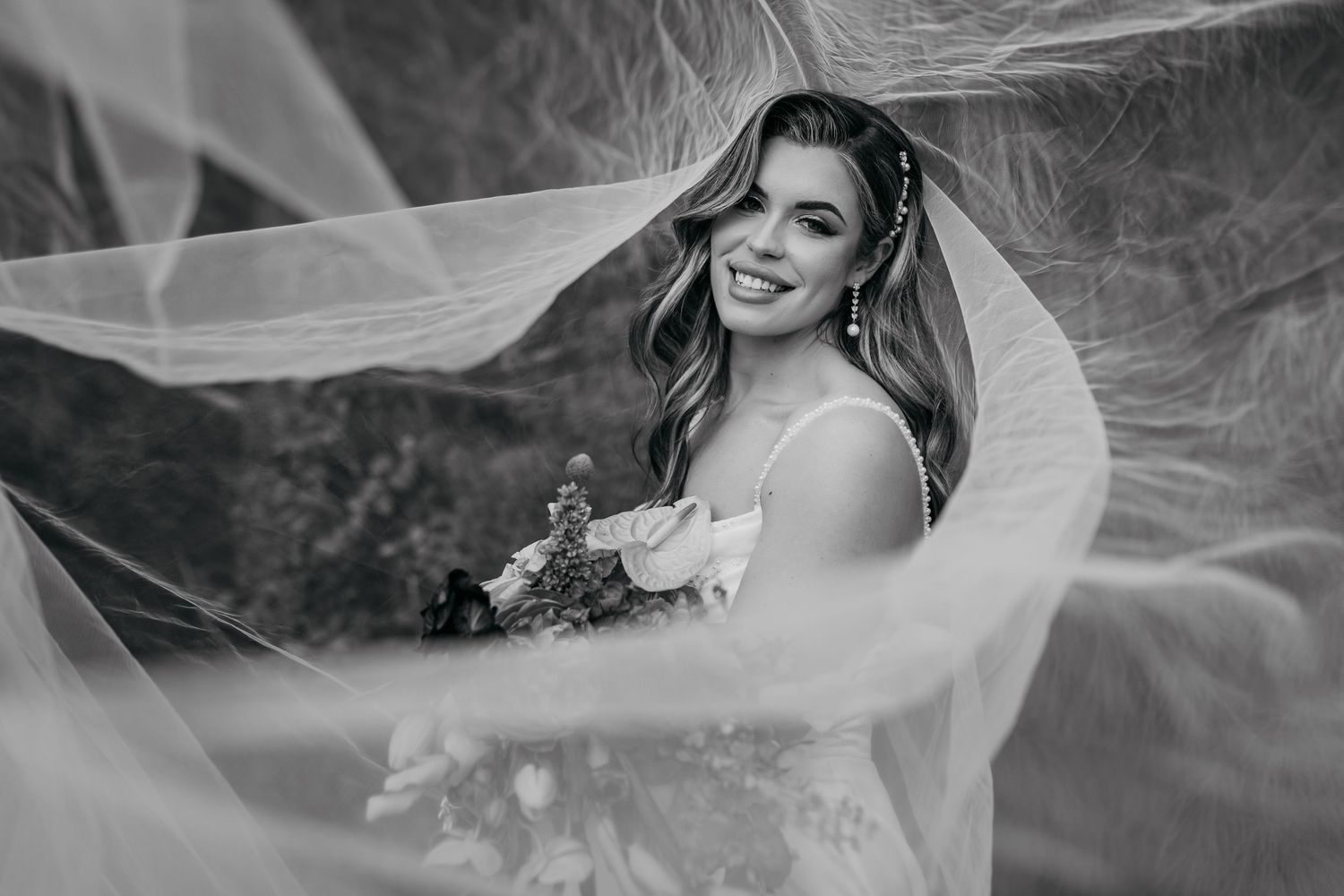 A black and white photo shows a bride in a flowing veil holding a bouquet during a romantic outdoor photoshoot.