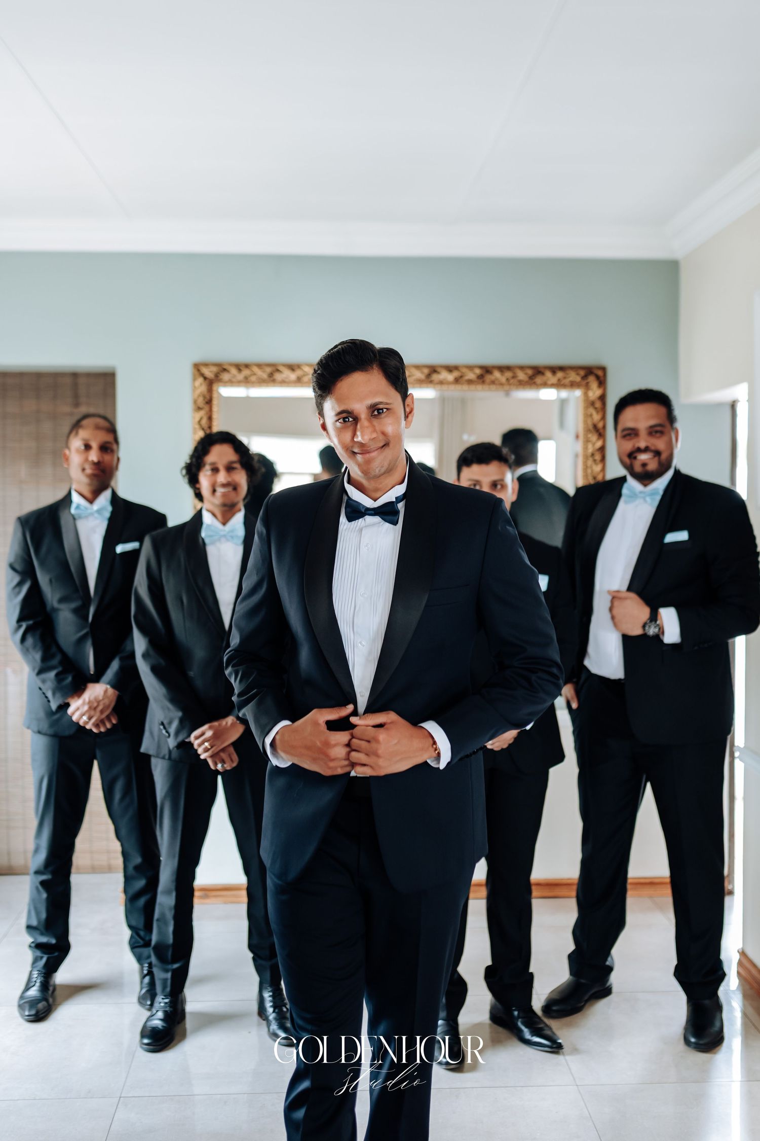 Groomsmen in black tuxedos and bow ties stand together in a bright room with a decorative mirror.