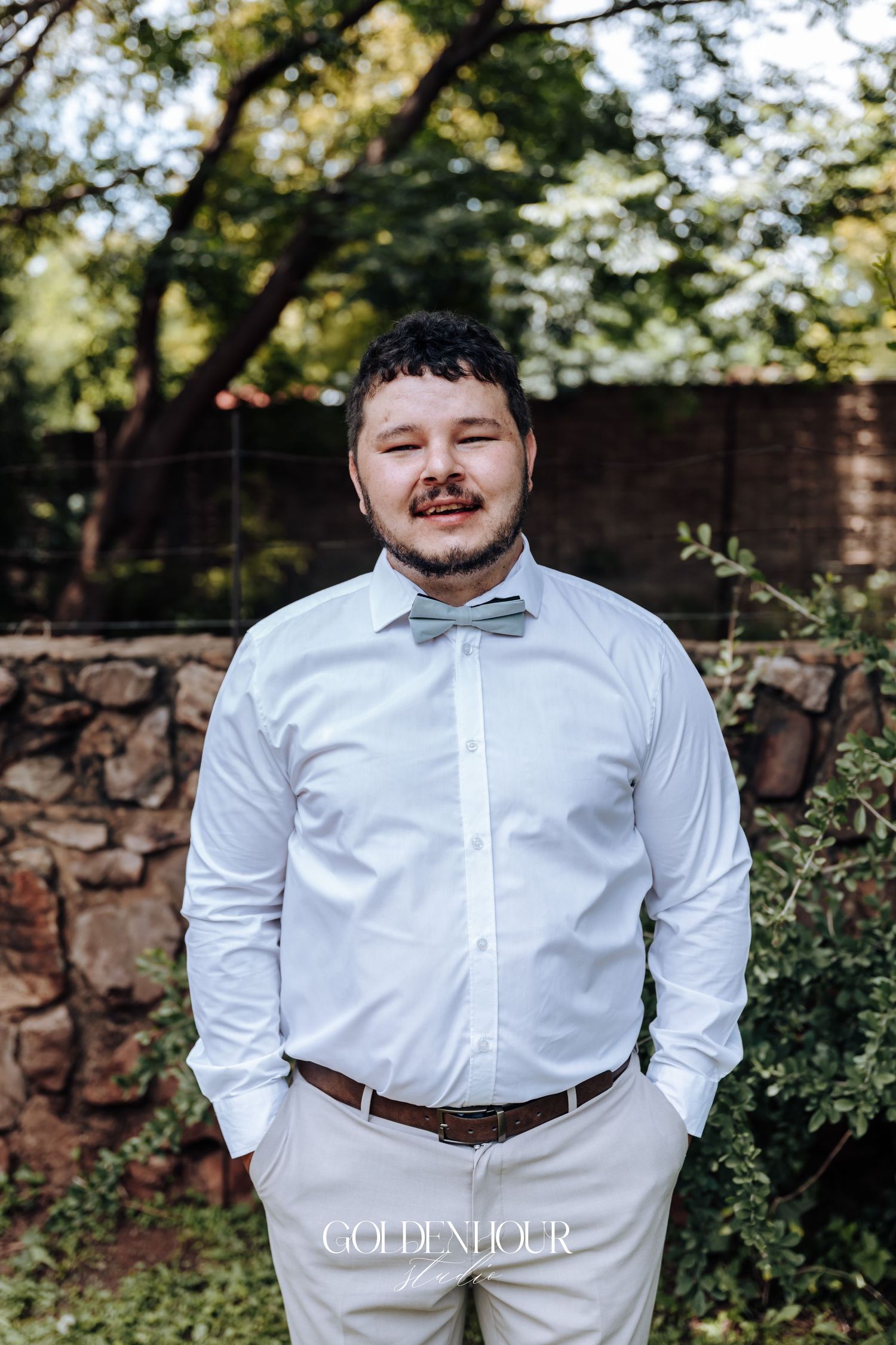 Formal attire portrait in white shirt and bow tie against stone wall and greenery backdrop.