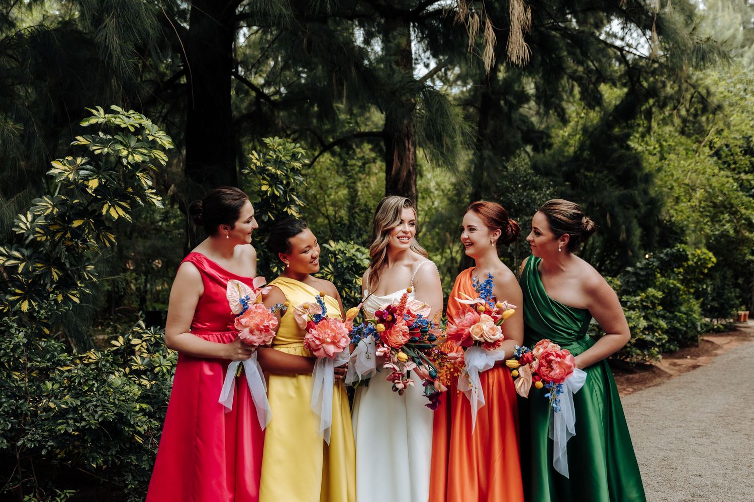 Bridesmaids wearing rainbow-colored dresses stand together holding colorful floral bouquets in a garden setting.