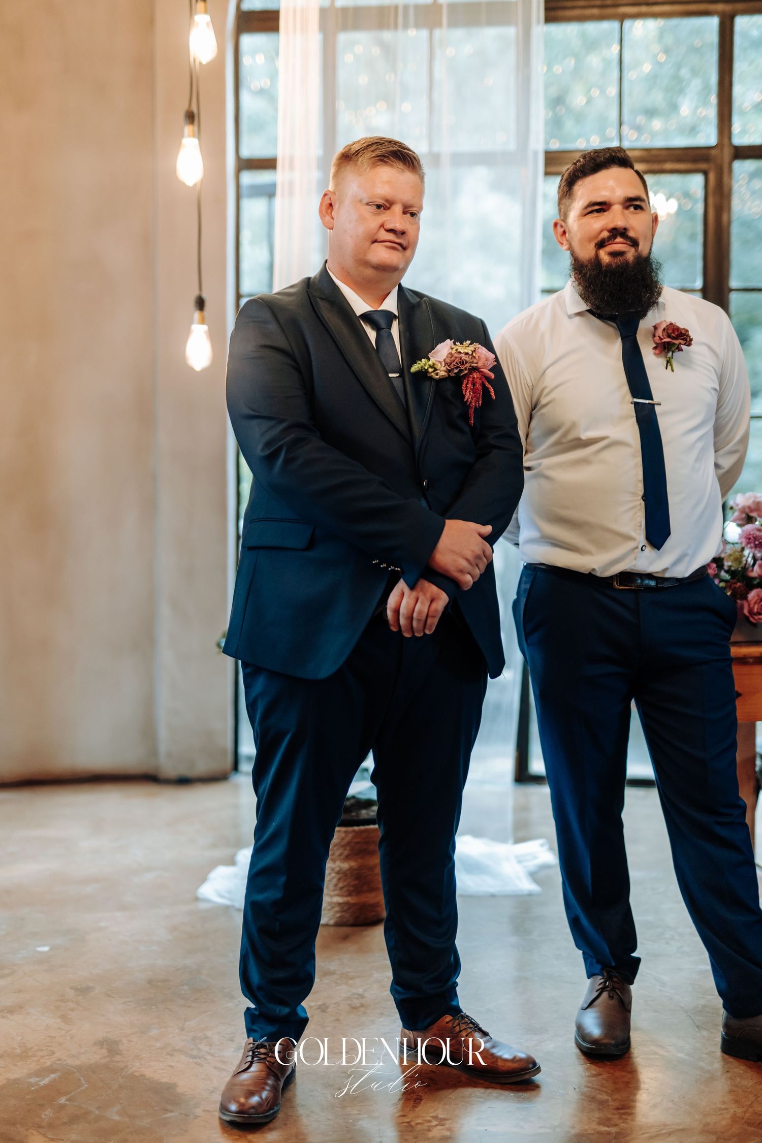 Two groomsmen in navy suits with floral boutonnieres standing at wedding altar in elegant industrial venue.