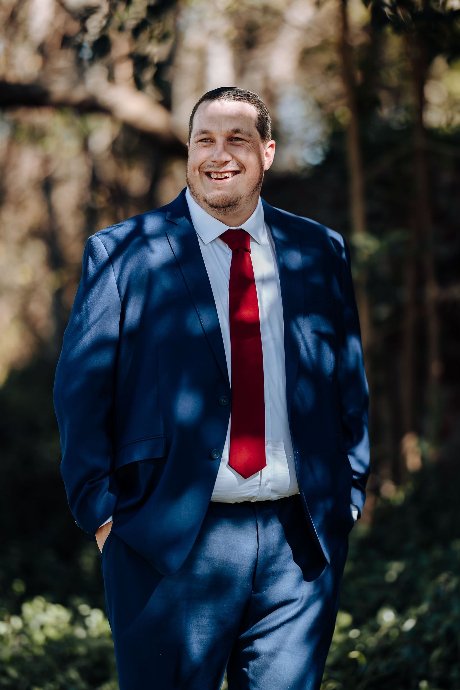 Smiling man in navy blue suit with red tie standing outdoors among green trees in a professional portrait.