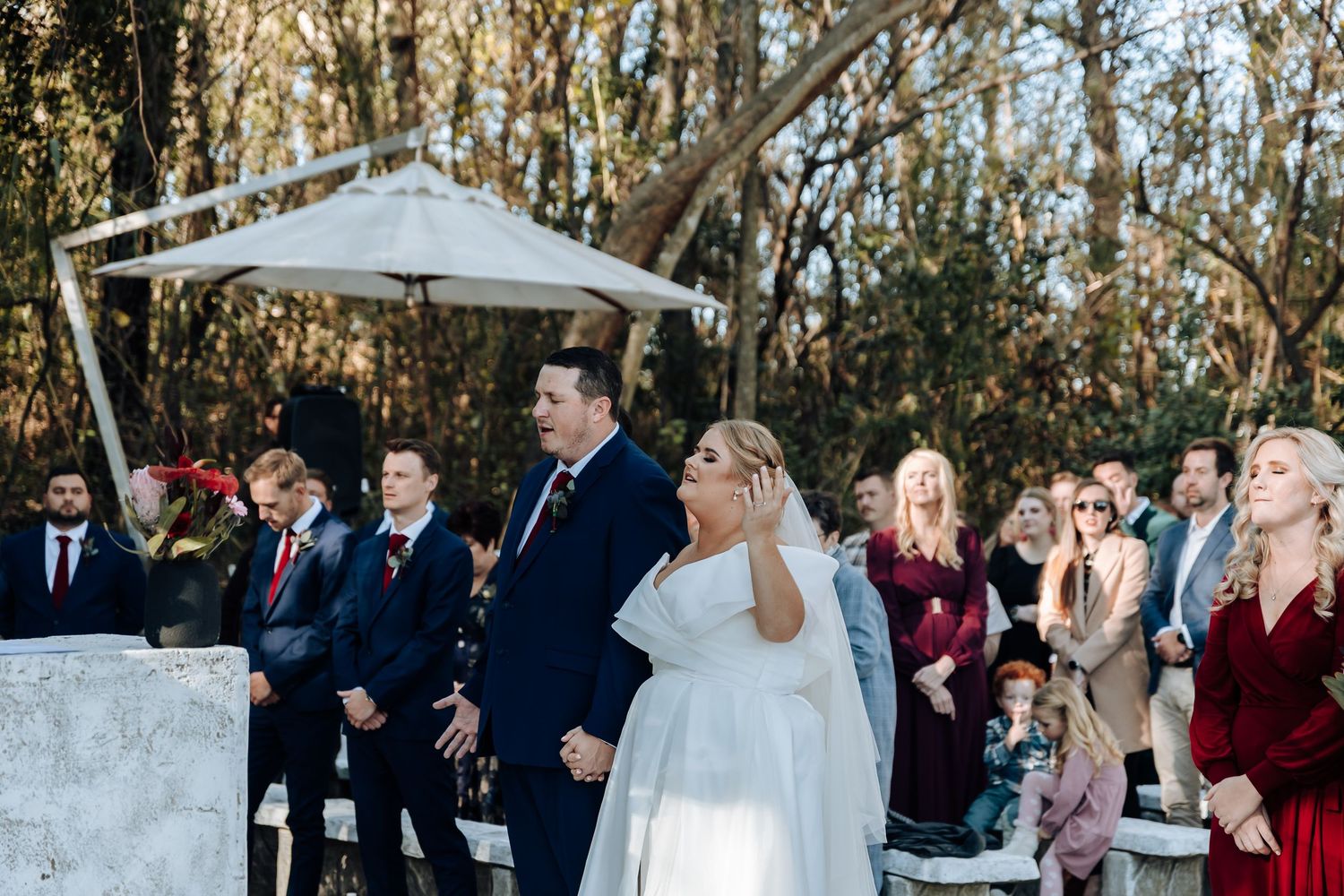 Bride in white gown walks down outdoor aisle with escort at elegant woodland wedding ceremony.