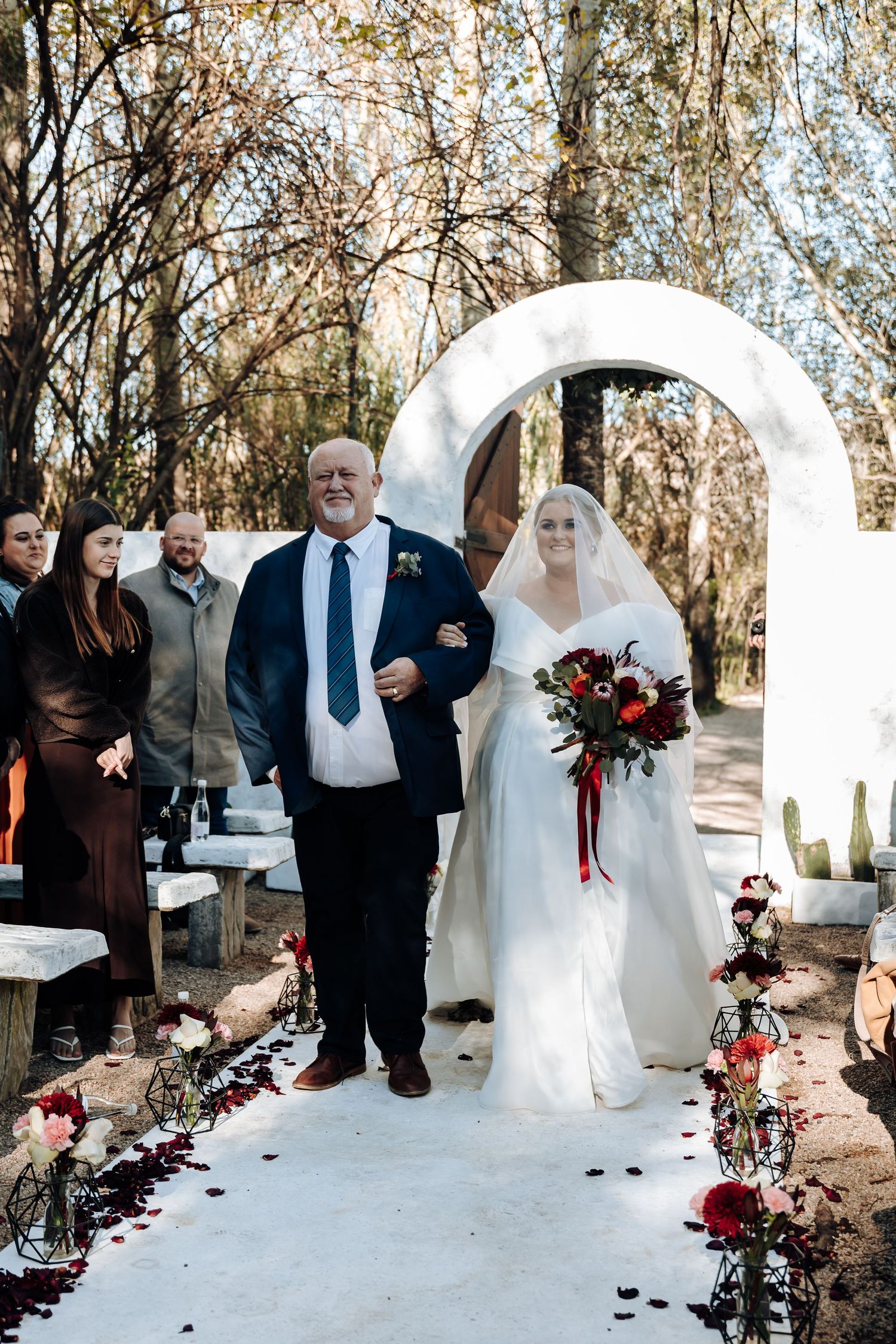 Bride walks down outdoor aisle with father under white arch, holding red bouquet, surrounded by autumn trees.