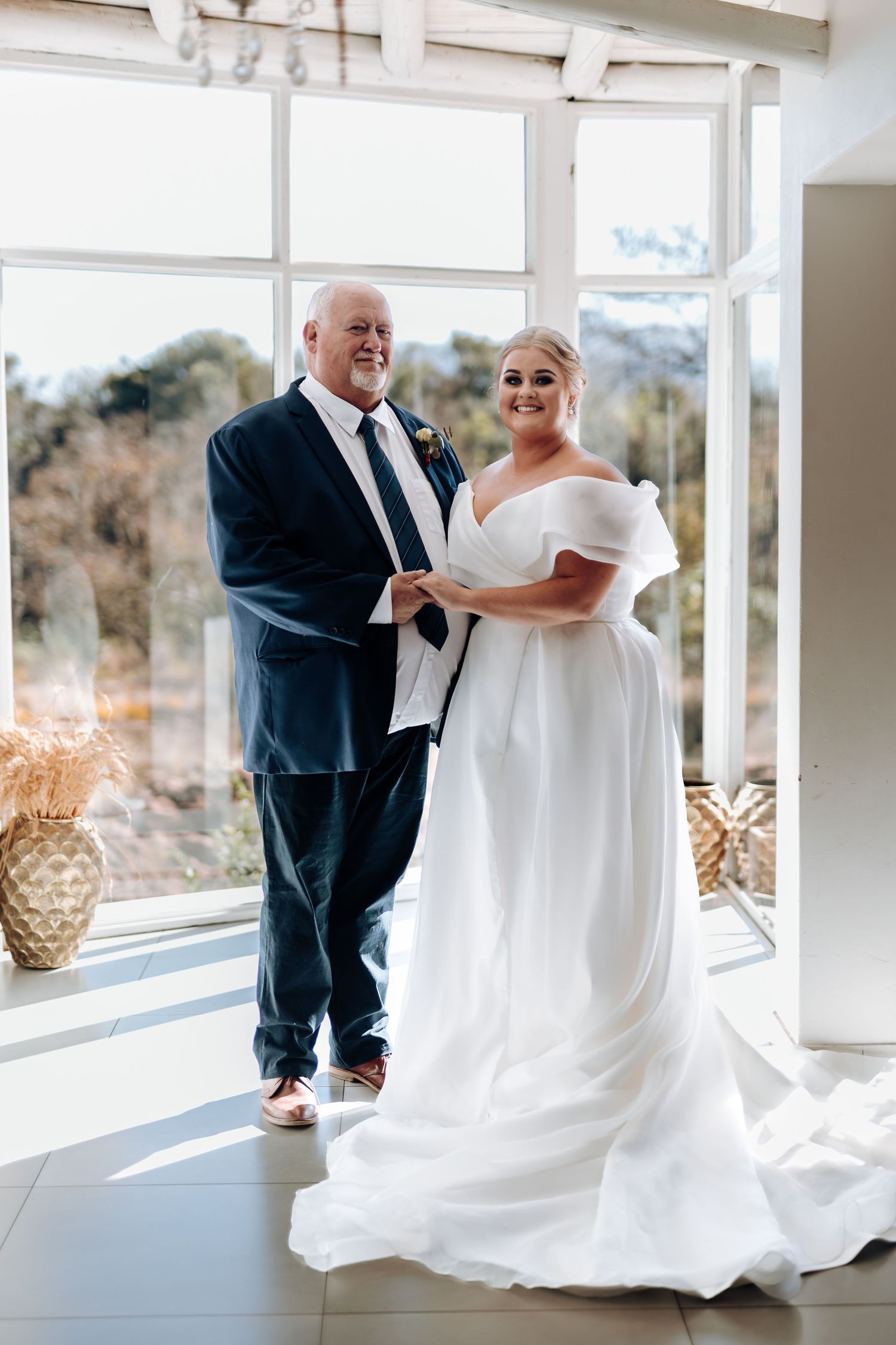 Bride in off-shoulder white gown and groom in navy suit pose together indoors on their wedding day.