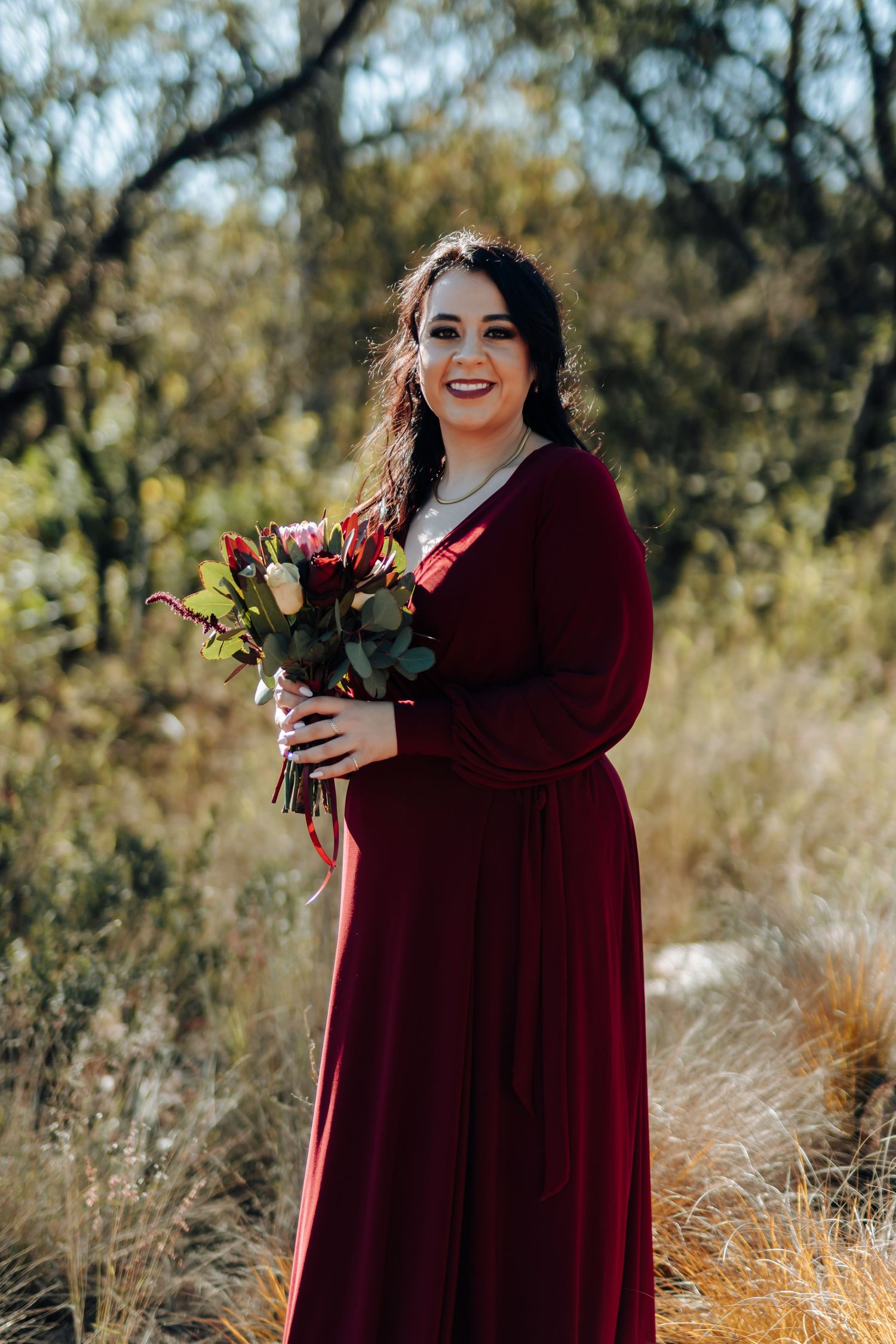 Woman in burgundy velvet gown holding floral bouquet smiling outdoors among autumn foliage.