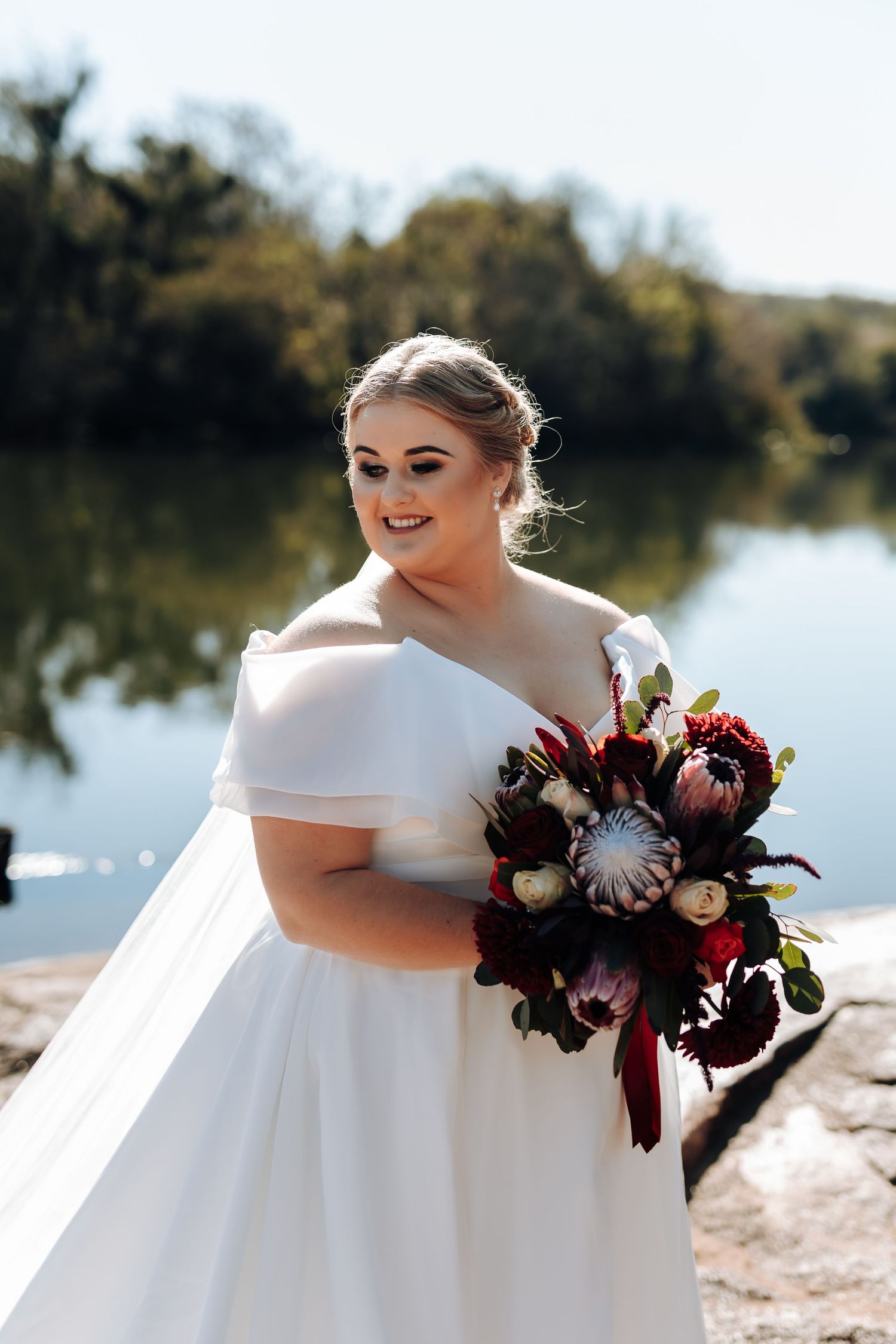 Bride in off-shoulder white gown holding a dark red bouquet with protea flowers, standing by a reflective lake.