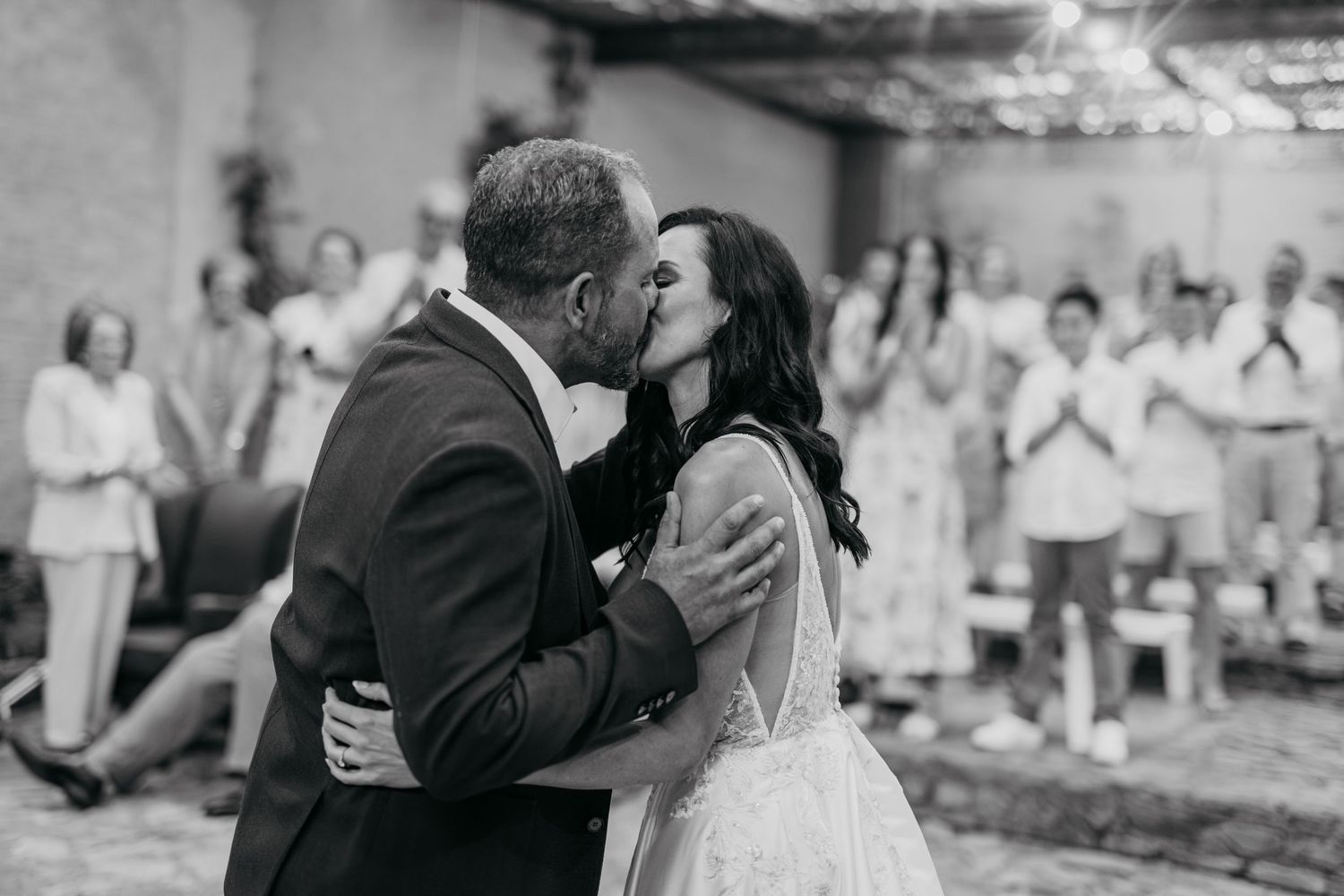 Black and white photo of a bride and groom sharing their first kiss at a wedding ceremony with guests applauding.